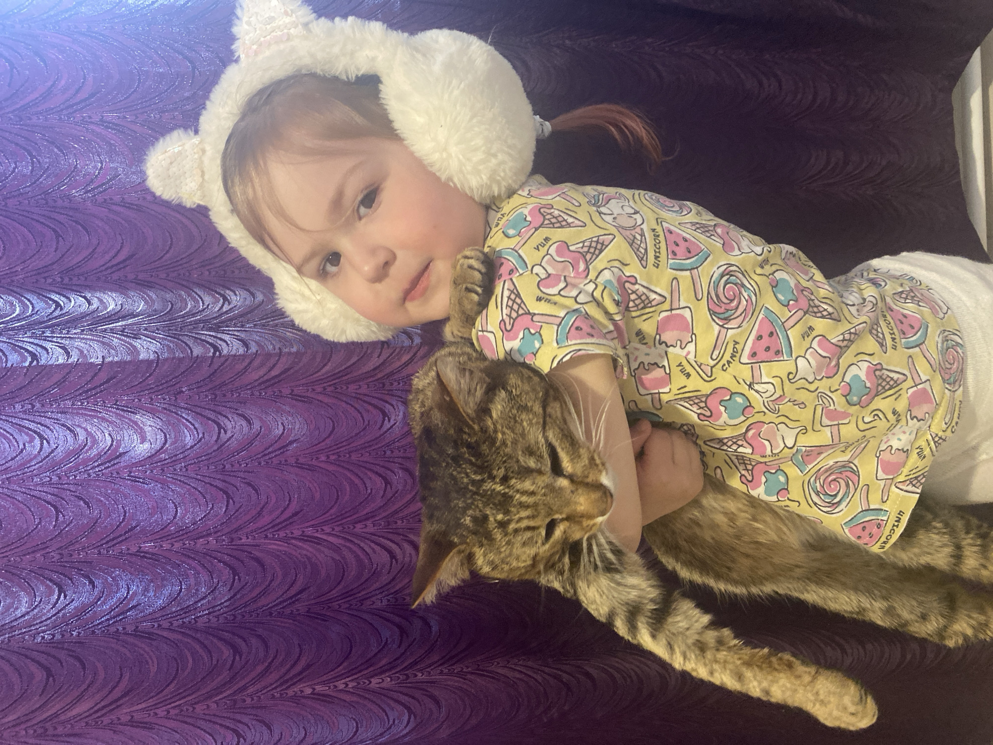 An overhead shot shows a young child and a tabby cat comfortably nestled together on a vibrant purple, textured surface. The child, likely a toddler or preschooler, lies on her left side, looking towards the viewer with a gentle expression. She has light brown hair styled in two small pigtails and is wearing fluffy white earmuffs that resemble animal ears, with subtle sequin details. Her short-sleeved yellow top is adorned with a playful pattern of pink and blue ice cream cones, popsicles, sliced watermelons, and lollipops, alongside visible text including "Yum", "Unicorn", and "Candy".
The cat, a brown and black tabby, is embraced by the child, its head resting near her left arm and chest. The child's left hand is wrapped around the cat's body, indicating a close cuddle. The cat appears relaxed, with its eyes partially closed. The background is a deeply textured purple fabric or blanket with an undulating, wave-like pattern that catches the light, creating highlights and shadows. The scene is lit, suggesting an indoor setting. The overall image conveys a sense of warmth, affection, and comfort between the child and her pet. This scene is set in Chaikovsky, Russia.