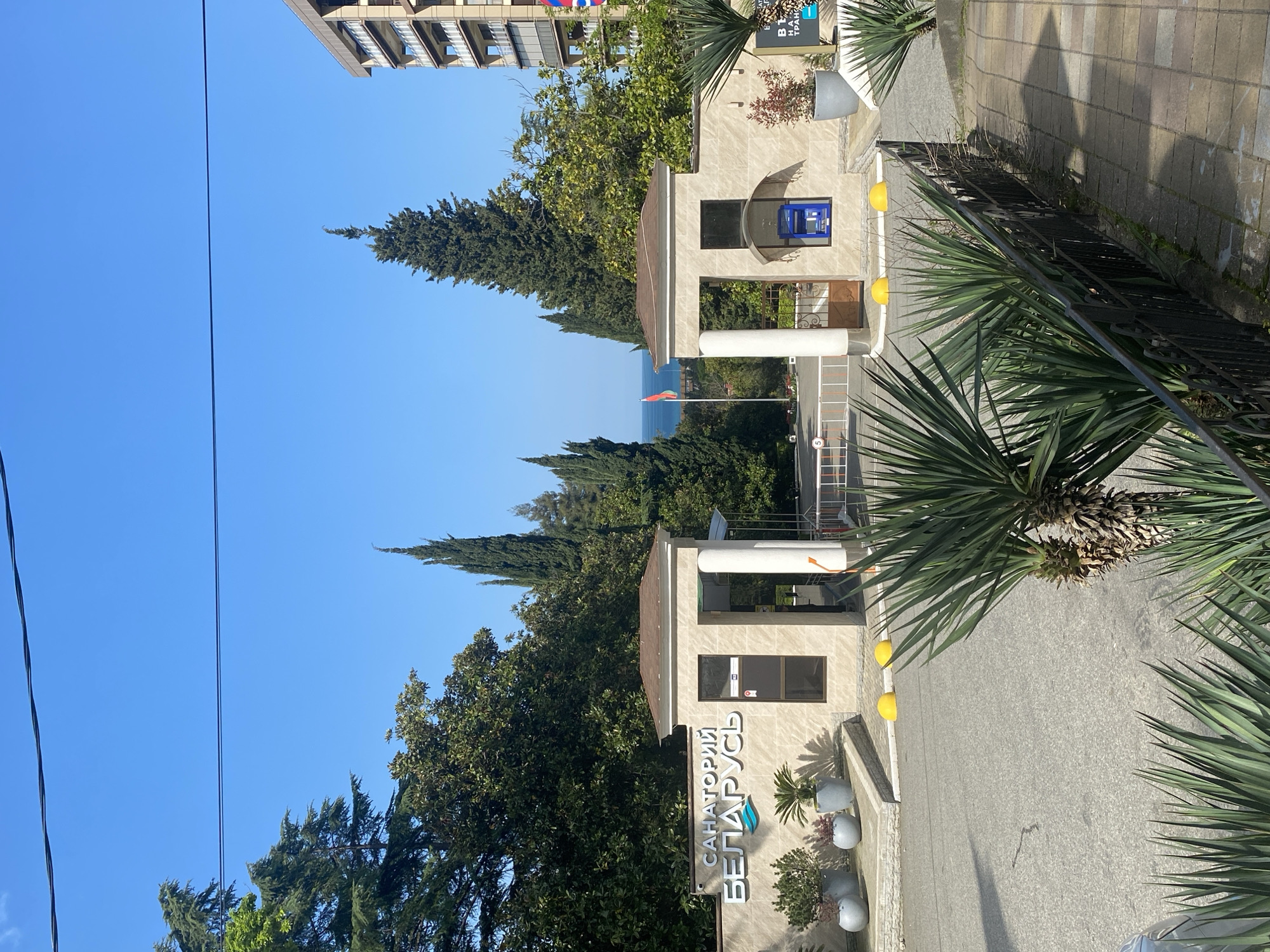The image captures the entrance to the "Sanatorium Belarus" in Sochi, Russia. The entrance consists of two matching gatehouses, each with a small window and a column. The building material appears to be a light-colored stone. A sign with the name of the sanatorium, "САНАТОРИЙ БЕЛАРУСЬ," is visible at the base of one gatehouse. An ATM is seen within one of the gatehouses.
Beyond the entrance, a road leads towards the sea, which is partially visible in the distance, as well as a Belarusian flag. Various trees and vegetation surround the area, including tall, thin conifers and palm trees. A building is visible in the upper portion of the image, to the right of the frame. An overhead wire runs through the frame, starting on the left side of the image. The weather appears sunny and clear.