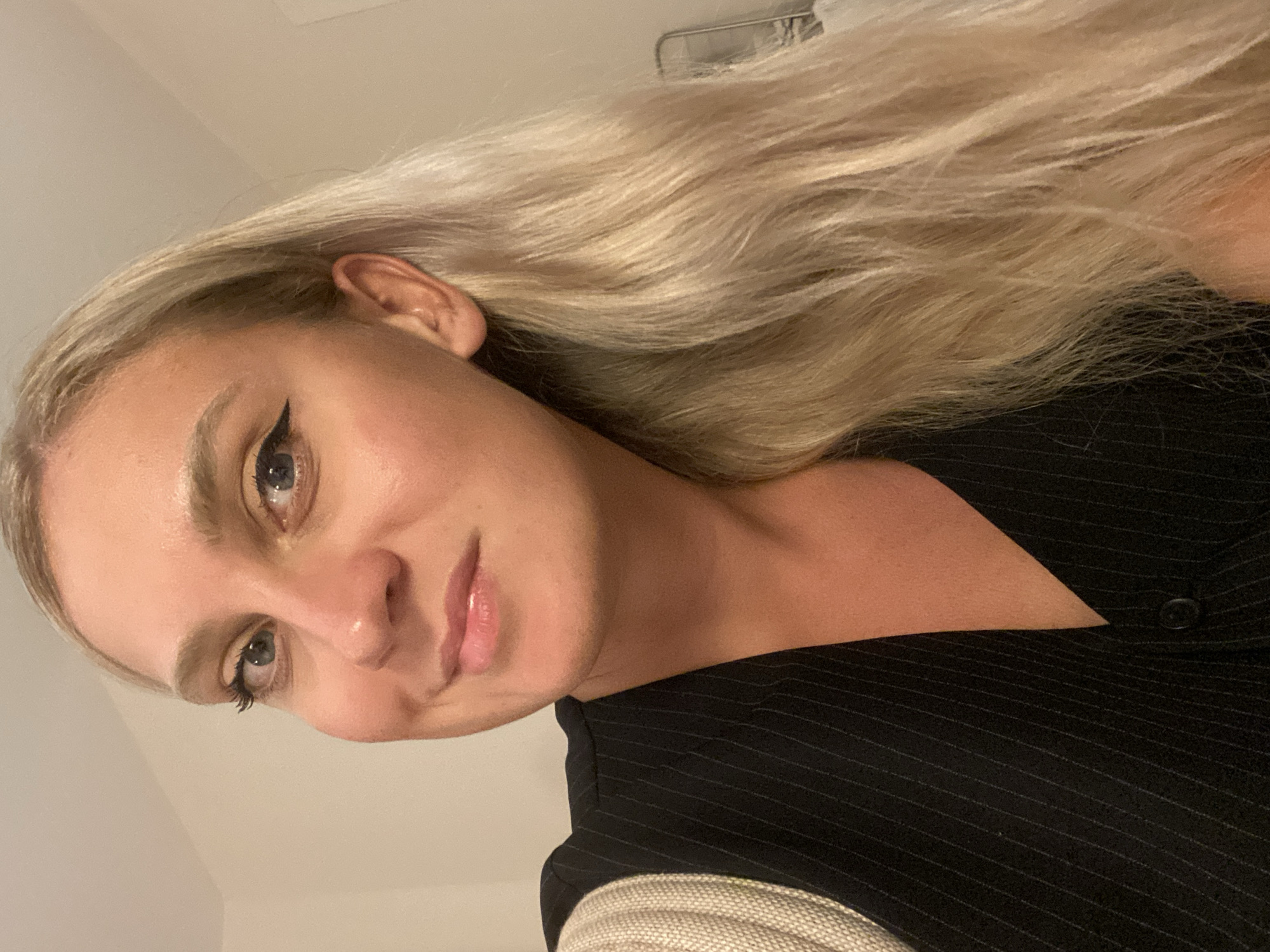 A young adult female with blonde hair and blue eyes is shown in a close-up portrait. She is wearing a black pinstripe vest over a beige ribbed top. Her makeup includes eyeliner and lip gloss. The background is a plain, light-colored wall. No specific location in Aksu, Turkey is indicated in the image.