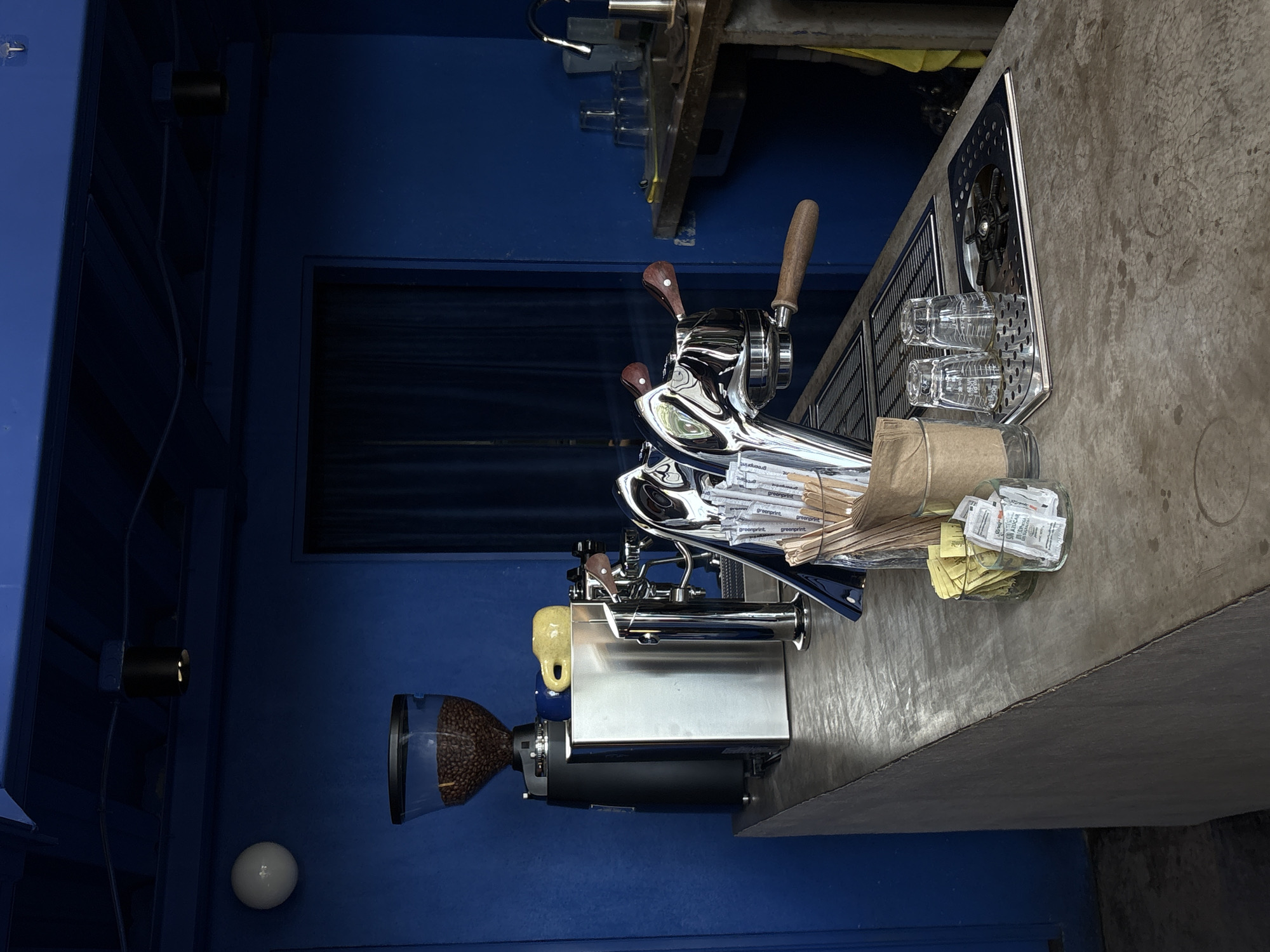 The image depicts a coffee counter in San Pedro Garza García, Mexico. The counter is made of concrete and is set against a vibrant blue wall. On the counter, there is a coffee grinder filled with beans, a small yellow mug, a chrome espresso machine, and various coffee-making accessories such as straws, wooden stirrers, sugar packets, and two empty glasses. To the right of the counter, a sink area is visible with stacked glasses and a faucet. The overall scene suggests a functional and well-equipped coffee preparation area.