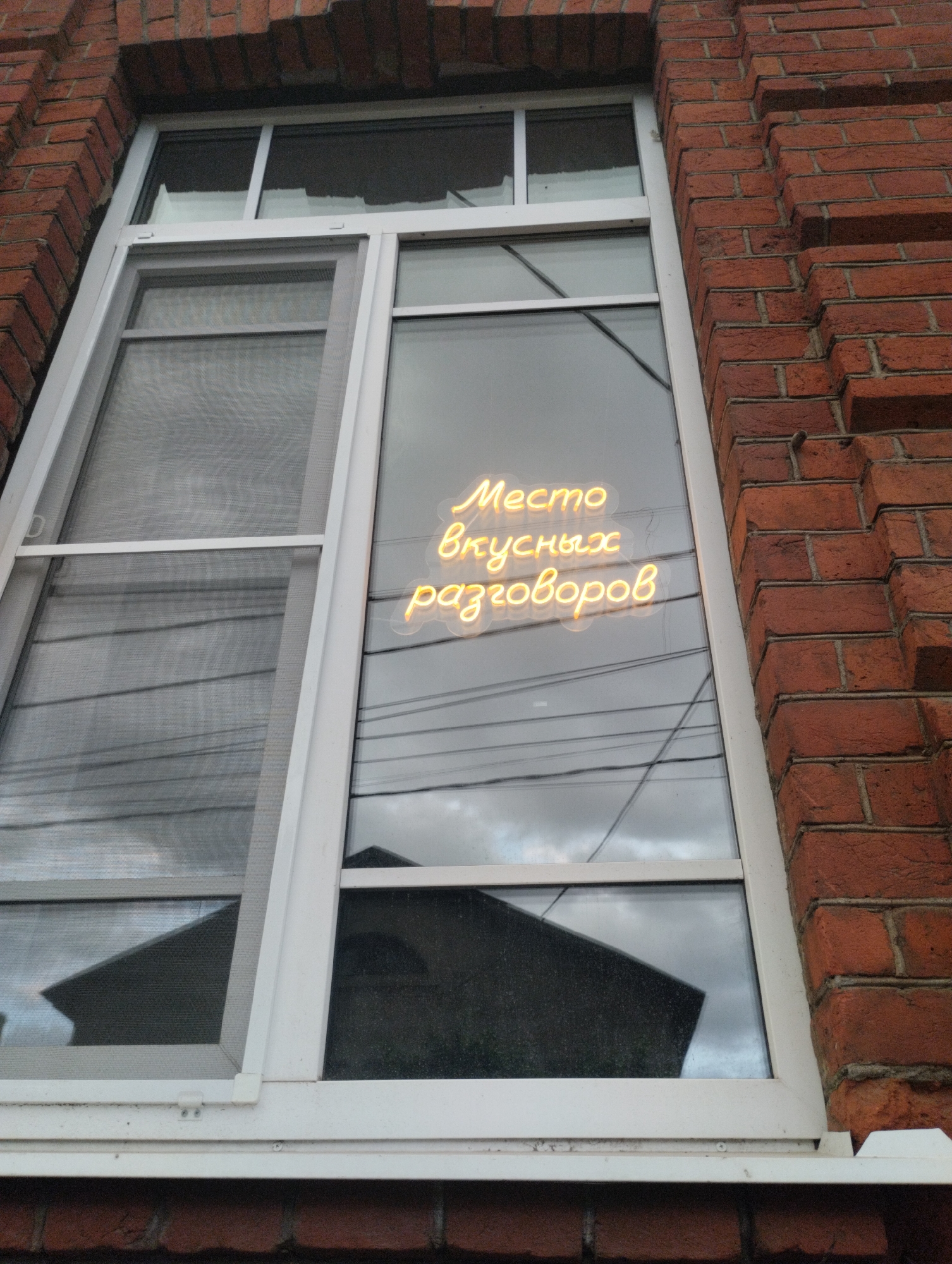 A close-up exterior shot shows a window on a brick building. The window is divided into two panes. The left pane is covered by a screen. The right pane displays a neon sign that reads "Место вкусных разговоров" (Place for tasty conversations). The sign is yellow and illuminated. Reflections of clouds and a dark roof are visible in the window. The building is made of red brick. The scene is a street-level view looking up at the window. This is likely a business with outdoor signage.