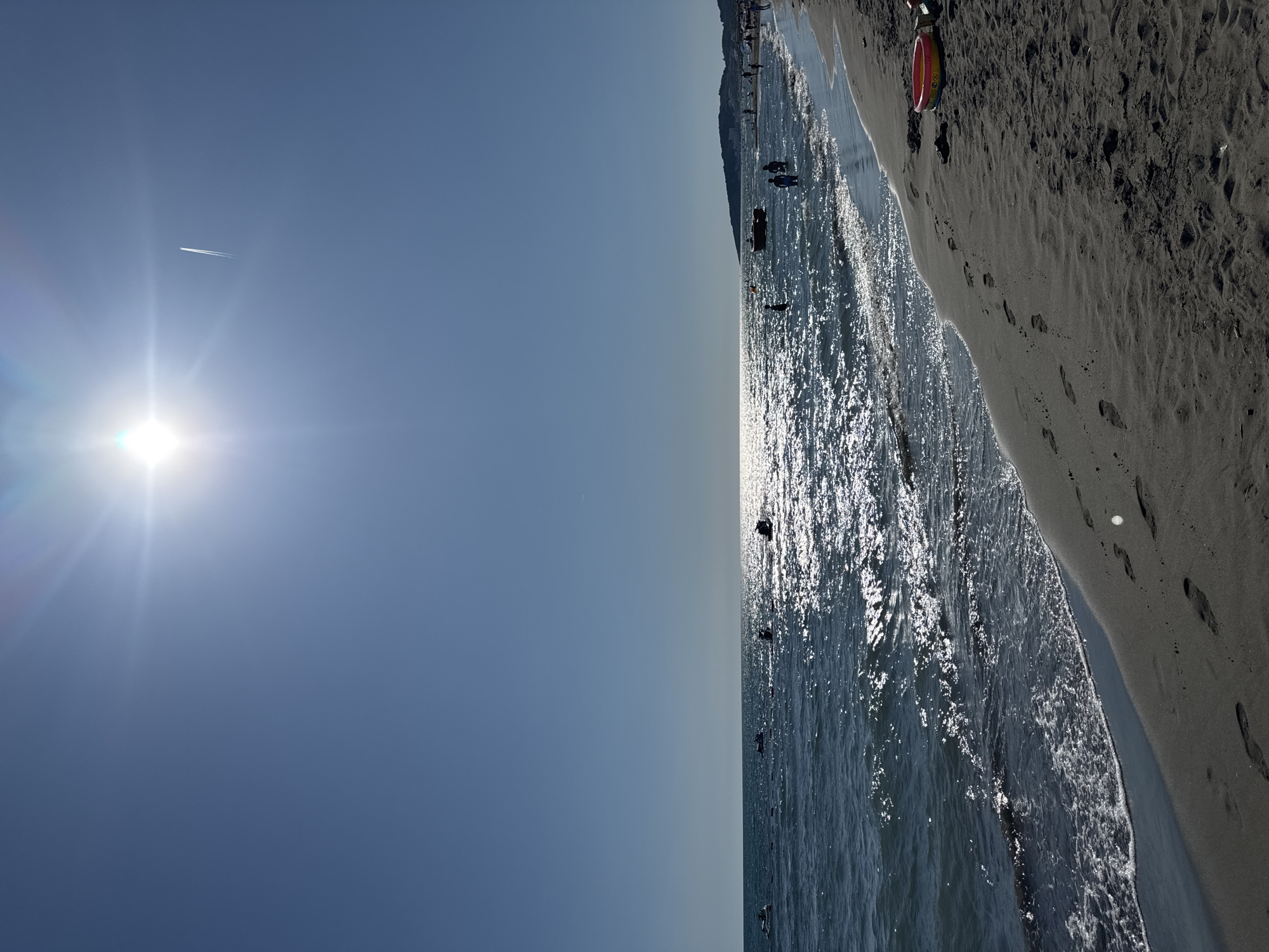 The image depicts a sunny day at a beach in Donji Štoj, Montenegro. The sky is clear blue with a bright sun and a contrail from an airplane. The sea is calm with gentle waves. There are several people in the water, some of whom appear to be on jet skis or small boats. The beach is sandy and there are footprints visible on the sand. A small colorful toy bucket is on the sand near the water's edge. In the distance, a hilly coastline is visible with some buildings. The overall scene is one of a pleasant summer day at the beach.
