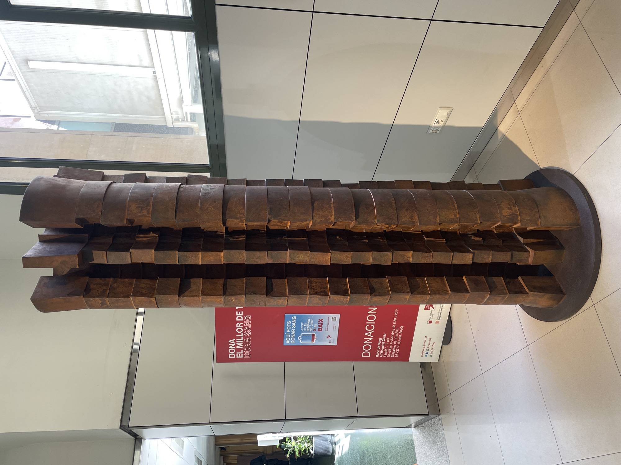 The image shows a tall, abstract metal sculpture in an indoor setting. The sculpture is brown and appears to be made of stacked, irregular rectangular blocks. To the left of the sculpture is a large red sign with white text. The sign includes information about donating blood and contact details for "Banc de Sang a l'Hospital Clinic." The floor is tiled with light-colored squares. In the background, through a window, there is a view of an outdoor area with some buildings. The environment suggests a public or institutional space, potentially a hospital or clinic lobby, given the blood donation sign. The sculpture itself is the central focus, positioned prominently within the frame.