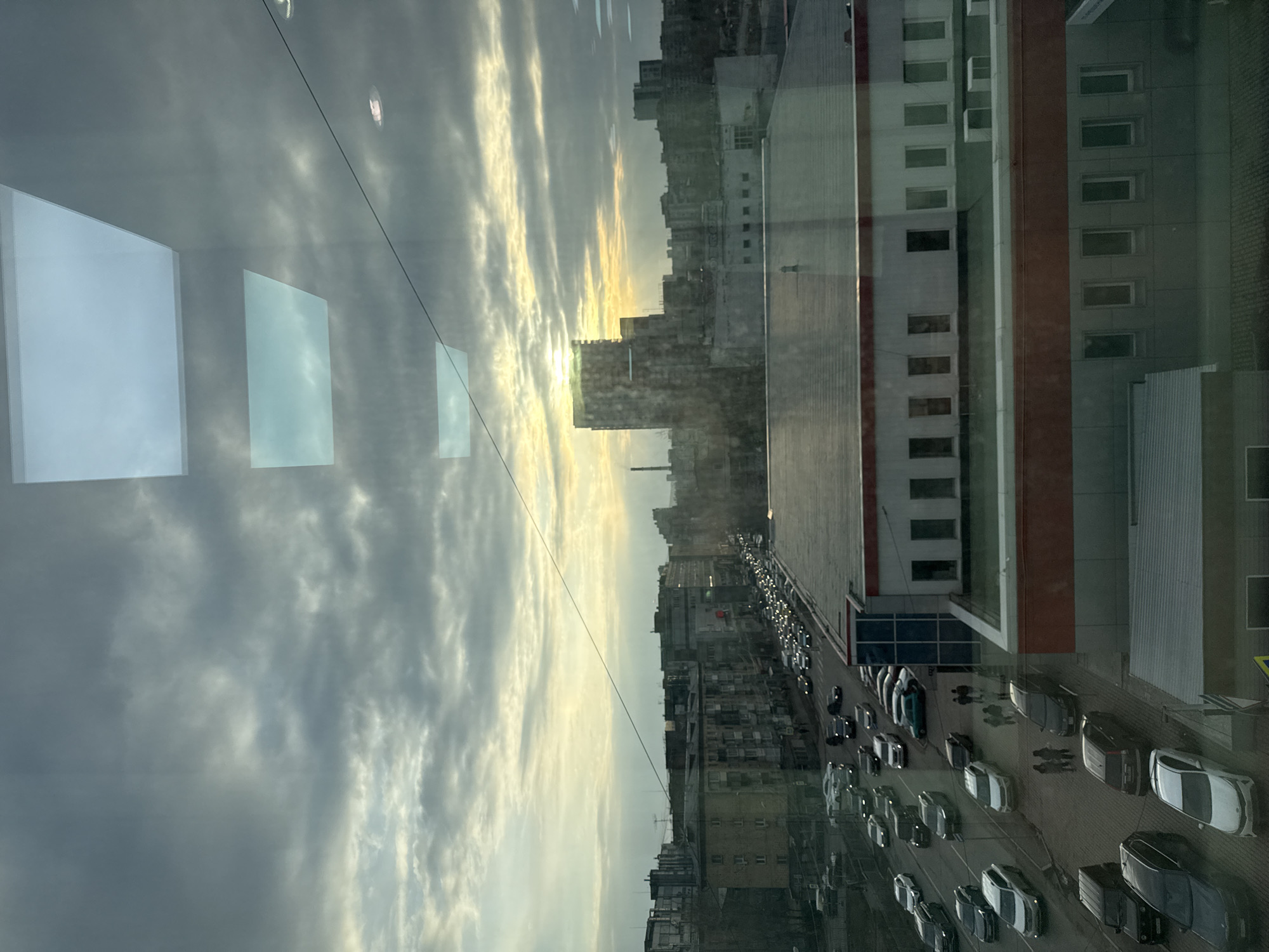 This is an upward-facing, indoor view of an urban scene, likely from a high-rise building in Krasnoyarsk, Russia. The scene shows a dense cityscape under a cloudy, overcast sky. The sun is partially obscured by buildings. The image is partly a reflection in glass. Reflected on the left side of the glass are some white rectangles, probably elements of the interior space. The skyline features buildings of varying heights, indicating a mixed urban development of old and new construction. A main street is visible with heavy car traffic, most of which are bright colored, and some pedestrians are on the sidewalks. To the right of the street is a large apartment or office building, and there are several other buildings along the street.