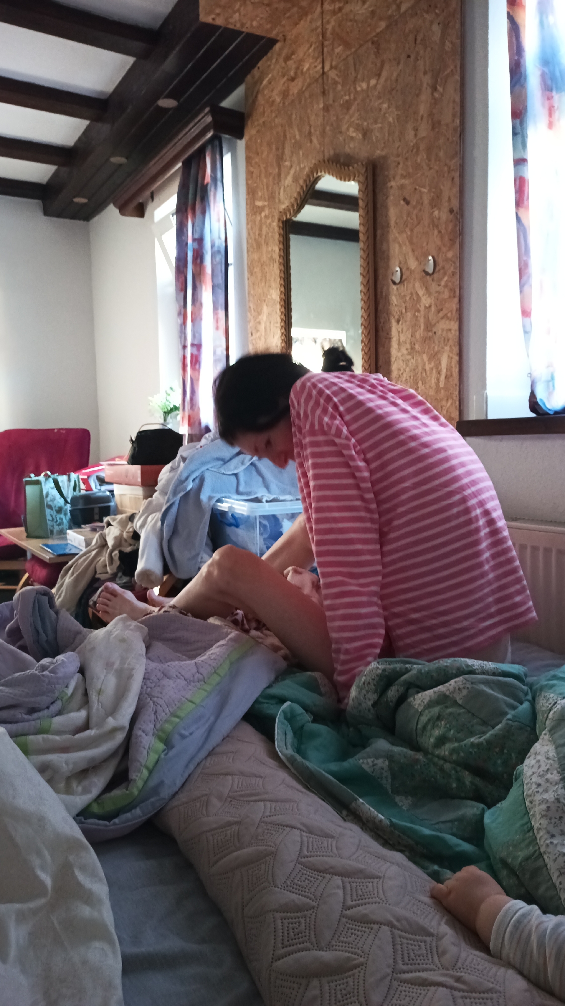 The image shows a woman in a pink and white striped long-sleeved shirt sitting on a bed. She is hunched over and appears to be looking at something in her lap or on the bed. Her legs are bent, and her right leg is extended forward. The bed is covered with a variety of blankets and pillows in different colors and textures. To the left of the bed, there is a red sofa, a wooden table with various objects on it including bags and books, and a window with patterned curtains. The room has a wooden beam ceiling and walls clad in wood panels. A mirror with a braided frame hangs on the wall. In the background, a radiator is visible. A young child's hand is visible in the lower right corner of the image, resting on a patterned pillow. The setting appears to be a bedroom or living space in Altdorf bei Nürnberg, Germany. The lighting suggests it is daytime.