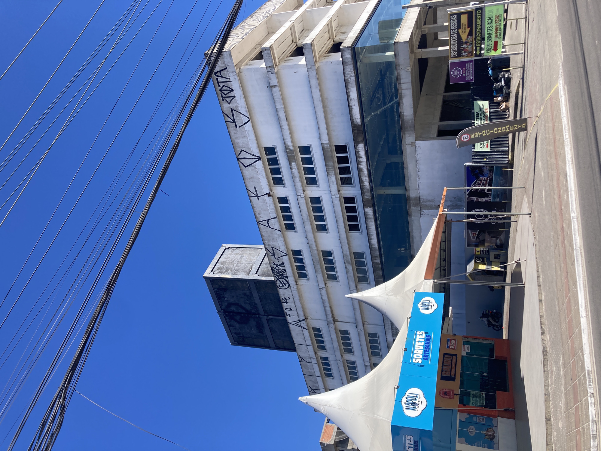 The image shows an urban scene in Florianópolis, Brazil. A tall, narrow building with graffiti on its side is visible. In front of the building is a covered outdoor space with advertisements for "Sorvetes Artesanais" (handmade ice cream) from Napoli Express. There are also signs for Distribuidora de Bebidas (beverage distributor), Estacionamento (parking), and Aiteria (pastry shop). Power lines are visible in the upper left corner of the image.
