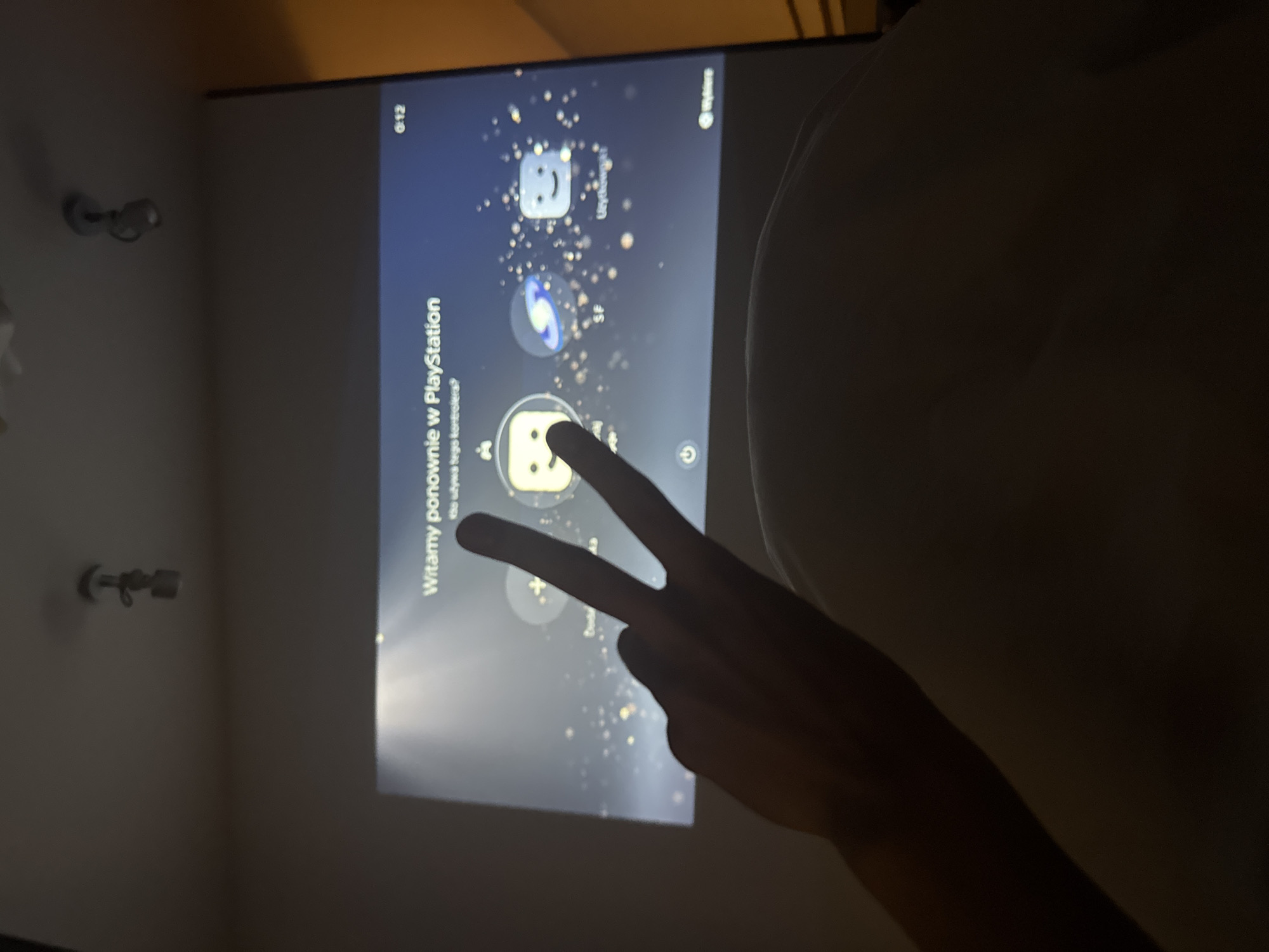 A slightly blurry, low-lit, medium shot depicts a television screen displaying a Playstation interface, intersected by a hand making a peace sign. The screen reads "Witamy ponownie w Playstation" with Polish text underneath. The time "0:12" is visible in the top left. The background is a neutral-colored wall, with electrical fixtures visible in the periphery. Location: Unknown. Warsaw, Poland is not discernable from the image.