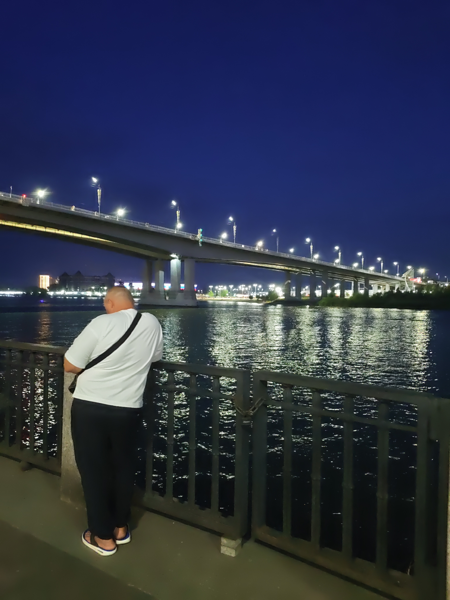 A man stands with his back to the camera, leaning against a dark metal railing. He is wearing a white t-shirt with a black strap across his back, and dark pants. The man is facing a wide river at night, with a large illuminated bridge spanning across it. The bridge is supported by concrete pillars and lined with streetlights. The water reflects the lights from the bridge and distant buildings. The sky is a deep, dark blue. The scene is likely the embankment in Rostov-on-Don, Russia, near the Voroshilovgradsky Bridge or the Don Bridge, given the presence of the river and a prominent bridge structure at night.
