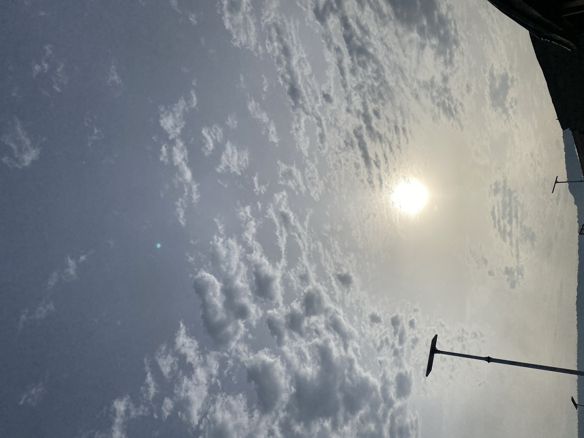 The image shows a cloudy sky with the sun visible through the clouds. The location appears to be Adankolo, Nigeria, based on the presence of utility poles. A building is visible at the top of the image.