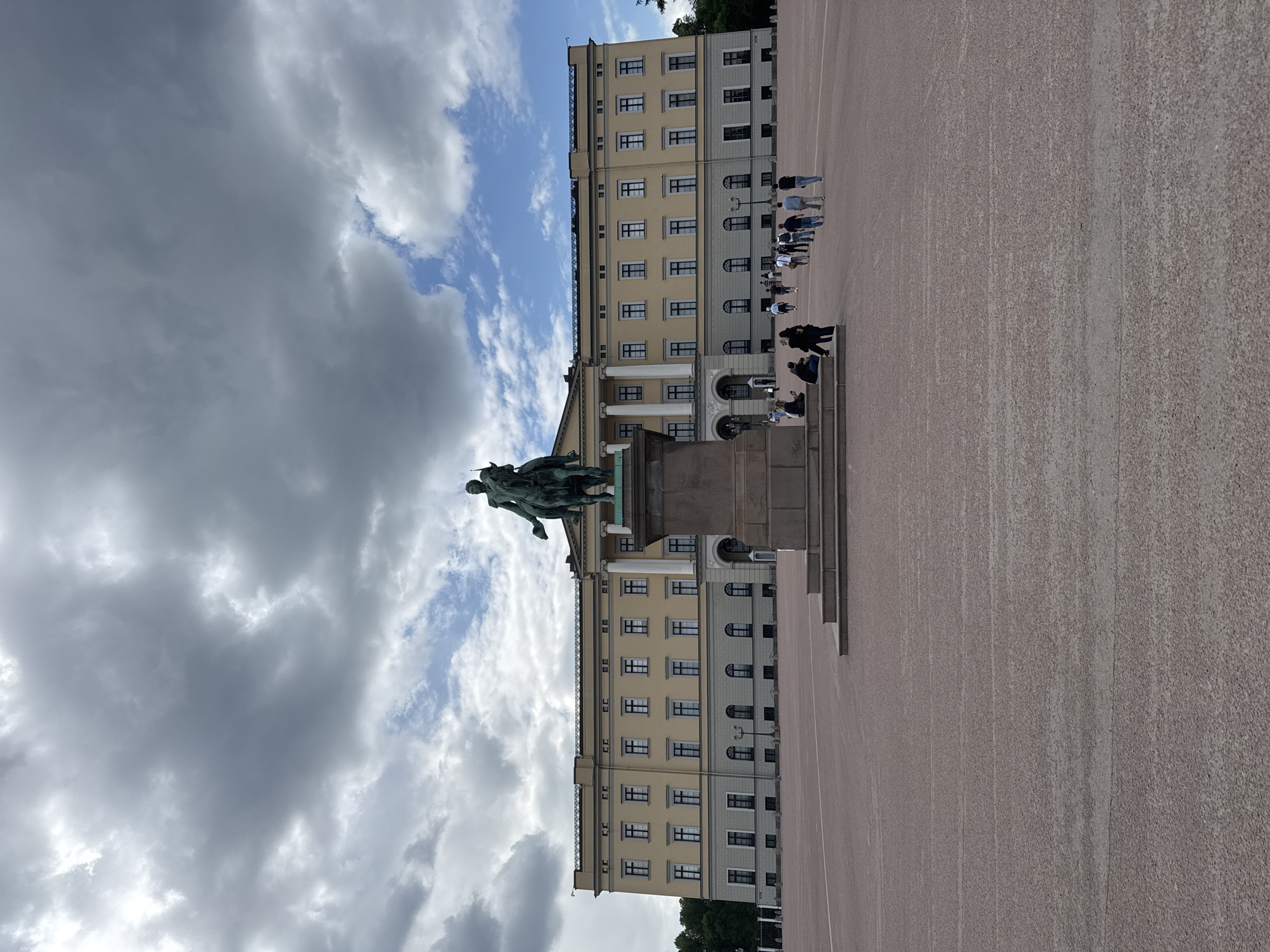 The image shows the Royal Palace in Oslo, Norway, with a statue of King Karl Johan XIV on horseback in the foreground. A large open space covered in fine gravel extends to the right of the palace. A group of people is standing in a line facing the palace, while others are seated on the steps leading up to the building. The palace itself is a multi-story building with a beige facade and dark-framed windows. The sky above is partially covered with dramatic clouds.