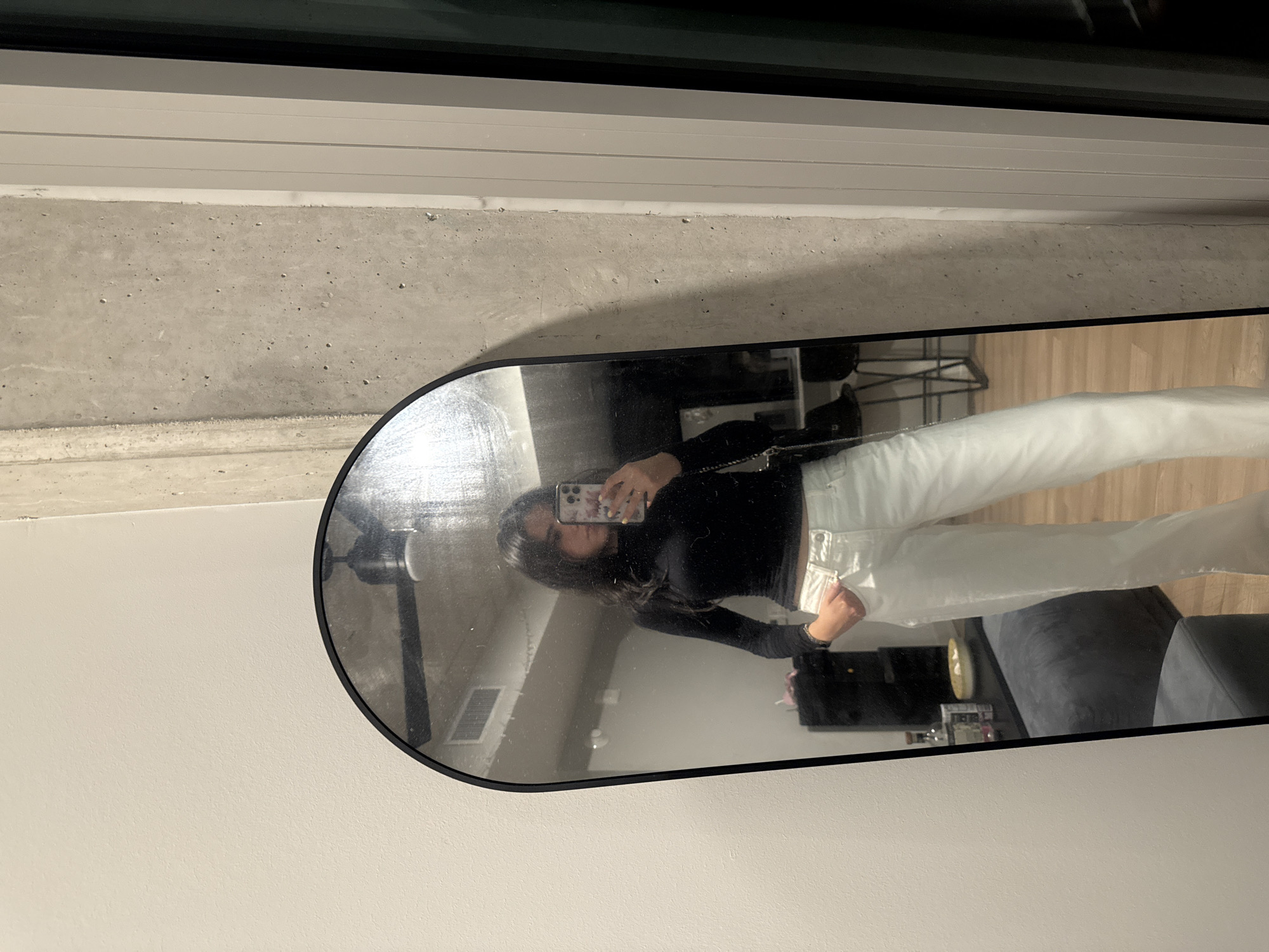 A vertical, eye-level indoor shot captures a person taking a mirror selfie. The image is oriented vertically with the top showing a dark window or door frame and a concrete wall section, transitioning to a light-colored painted wall on the left.
The main subject is a young person, likely female, with dark hair, visible from the chest up in the reflection of a large, rounded rectangular mirror with a black frame. She is wearing a long-sleeved black top and light-colored (possibly white or light blue) pants. Her right arm is raised, holding a smartphone with a distinctive patterned or sticker-covered case, as she uses it to capture her reflection. Her expression is neutral, focused on the phone.
The mirror's reflection reveals more of the room. To the person's right, a dark gray sofa or sectional is visible. Further into the reflection, on a surface behind the sofa, there are several bottles and possibly a decorative item. A silver rectangular vent is visible on the wall behind the person's head in the reflection. The floor in the reflection appears to be light-colored wood. The concrete wall texture and a light-colored horizontal window or door frame are visible above the mirror in the main image.
The lighting suggests an indoor setting, likely artificial or indirect natural light. There is no clear indication of the time of day or weather. The overall environment appears to be a modern residential or commercial space, possibly in Austin, United States of America, given the location context. No distinct text is visible apart from the indistinct pattern on the phone case.