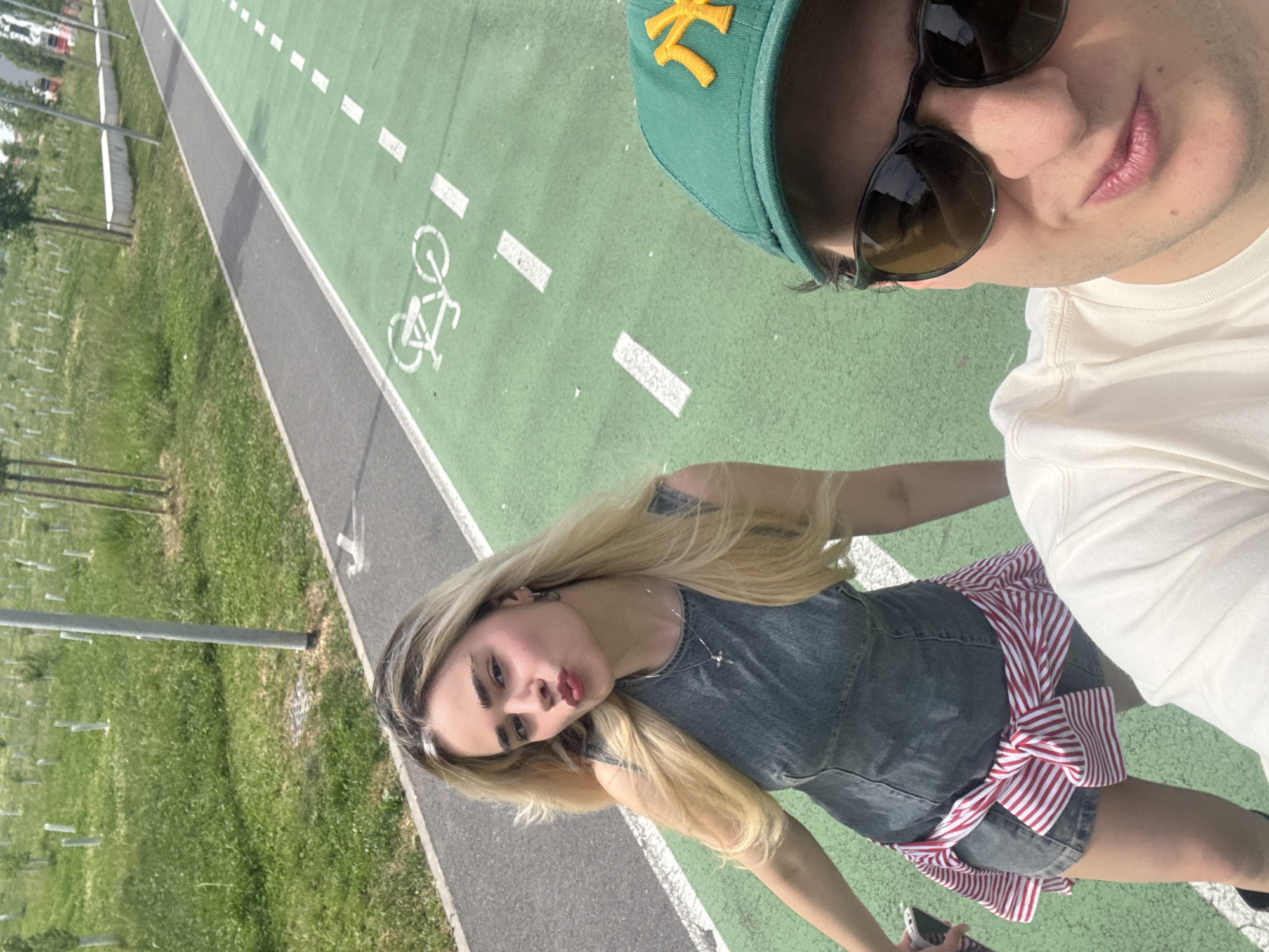 Two individuals pose for a selfie on a pedestrian and bike path. One wears a green cap with a yellow logo, sunglasses, and a white shirt. The other wears a denim dress with a red and white striped sash and has blonde hair. The path is marked with white dashed lines and a bicycle symbol. The environment consists of grass and some small trees. The photo was likely taken in Milan, Italy.