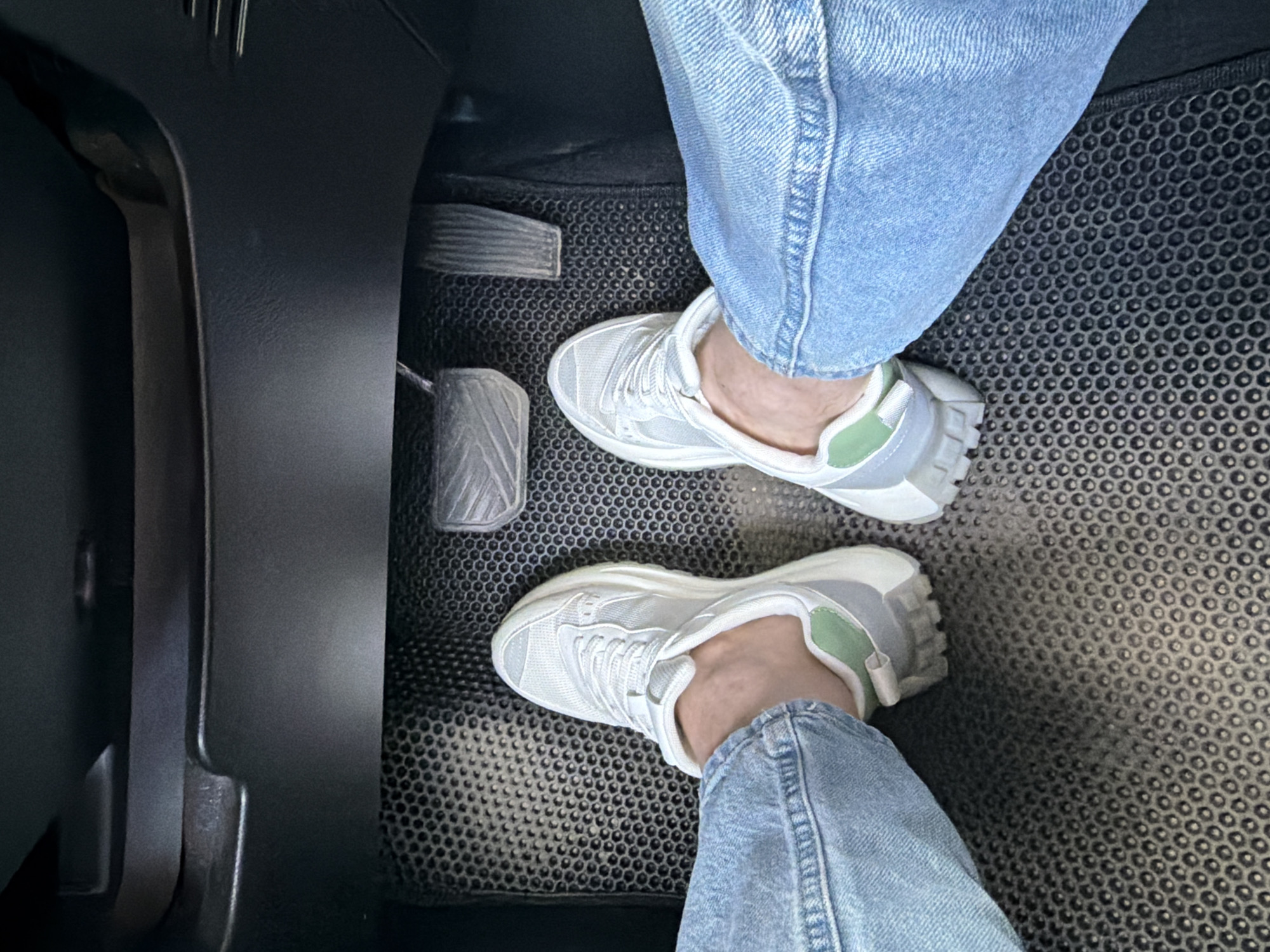 In the interior of a vehicle, likely a car, the perspective is low, focusing on the floor area near the pedals. Two feet, clad in light blue denim jeans and white sneakers with light green accents, are positioned on a black, hexagonal-patterned floor mat. The sneakers appear to be athletic or casual in style. The floor mat covers the lower portion of the vehicle's interior, extending towards the driver's area. Visible above the feet are three metal pedals: a clutch pedal, a brake pedal, and an accelerator pedal, arranged in the typical configuration for a manual transmission vehicle. The pedals are worn and appear to be made of metal. The surrounding environment consists of dark, molded plastic panels typical of a car's dashboard and footwell. The lighting suggests an indoor or shaded environment, possibly during the day, as there are no strong shadows or direct sunlight. The scene is static, showing the feet at rest on the floor. There is no indication of the specific location within the city of Chaikovsky, Russia, beyond the assumption that this is a vehicle interior.