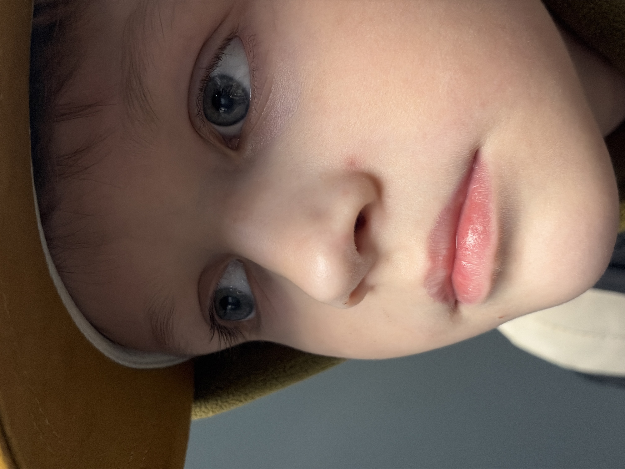 A close-up, slightly rotated shot focuses on the face of a young child. The child has pale skin, gray eyes, and light-colored hair partially visible at the top of the frame. The right side of the face is in soft focus, highlighting the eye and nose. A portion of a yellow or mustard-colored object, possibly clothing or a hat, appears in the lower left corner of the frame. The background is a muted gray or blue. The location is not discernible from the provided image.