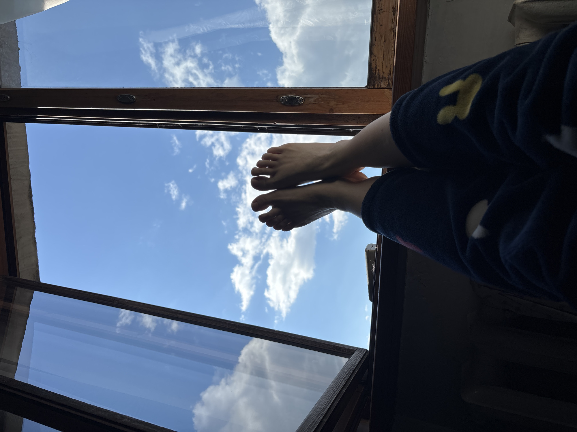 A low-angle, close-up shot focuses on a person's crossed bare feet extending from a window opening, with a view of a blue sky with scattered clouds visible through the window. The window is framed by dark, worn wood. The person is wearing dark blue, possibly fleece, pajama pants or pants with yellow star-shaped designs on the right pant leg. The window is slightly ajar, revealing more of the sky and cloudscape. The location and city of Unknown, Russia are not identifiable in the image.