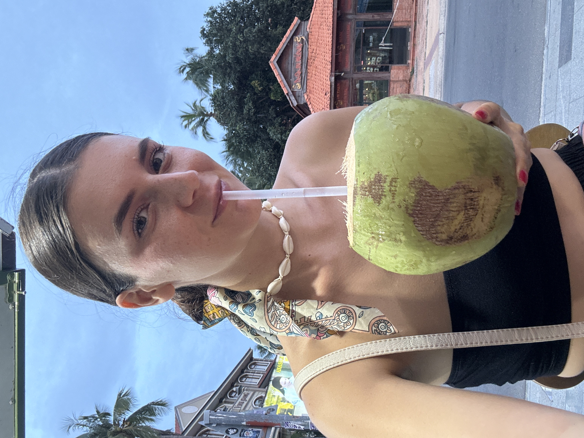A young woman is taking a selfie outdoors in Phu Thuy Ward, Vietnam, while drinking from a fresh coconut with a straw. She is wearing a black strapless top, a patterned scarf tied around her ponytail, and a necklace made of seashells. The background shows a street scene with buildings, palm trees, and a cloudy sky. One building has a red tiled roof and an ornate sign.