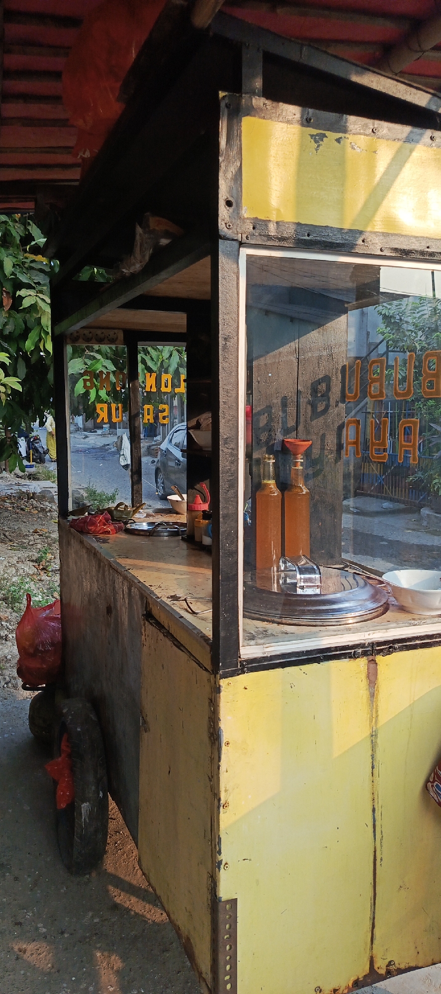 A street food cart, painted yellow and with a weathered appearance, sits on a paved surface, likely a street or roadside in Sukaresmi, Indonesia. The cart has a glass display area on the right side, revealing two bottles of amber liquid, a silver lid for a pot, and a white bowl. The words "BUBUR AYAM" are visible in orange lettering on the glass, indicating it sells chicken porridge. Through the open section of the cart on the left, a street scene is visible with a few trees, a parked silver car, and a person in the background wearing a yellow and white patterned outfit. The cart is equipped with a wheel on the lower left side, with a red plastic bag tied to it. The setting sun casts long shadows, suggesting it is late afternoon or early evening.