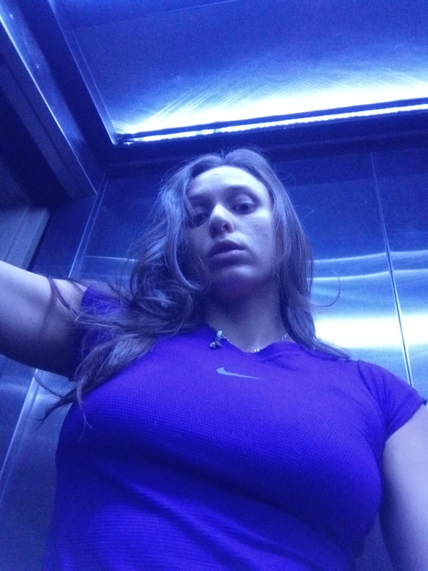 A woman is pictured in a metallic elevator under bright, bluish light. She is wearing a purple t-shirt with a Nike logo. The interior of the elevator is visible, featuring metallic walls and an illuminated ceiling panel. The location within Buenos Aires, Argentina is indiscernible.