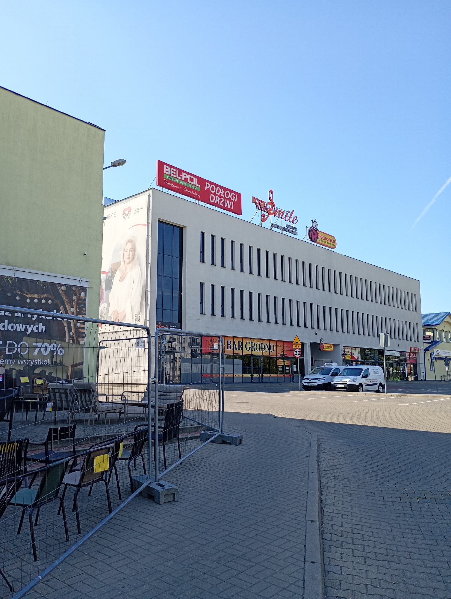 The image shows a modern building with a light-colored facade and dark windows, situated in Białystok, Poland. The building features prominent signage, including "BEL-POL PODŁOGI DRZWI" and "Your smile," suggesting it houses retail establishments. Several shops are visible on the ground floor, including "BAR GRODNO" and "Biedronka." Two white vans are parked in front of the building. In the foreground, there is a temporary fence enclosing an area with chairs and tables, likely an outdoor seating area for a restaurant or cafe. A large billboard with an image of a woman and promotional text is visible on the side of an adjacent building. The sky is clear blue, indicating a sunny day. The street in front of the building is paved with gray interlocking stones.