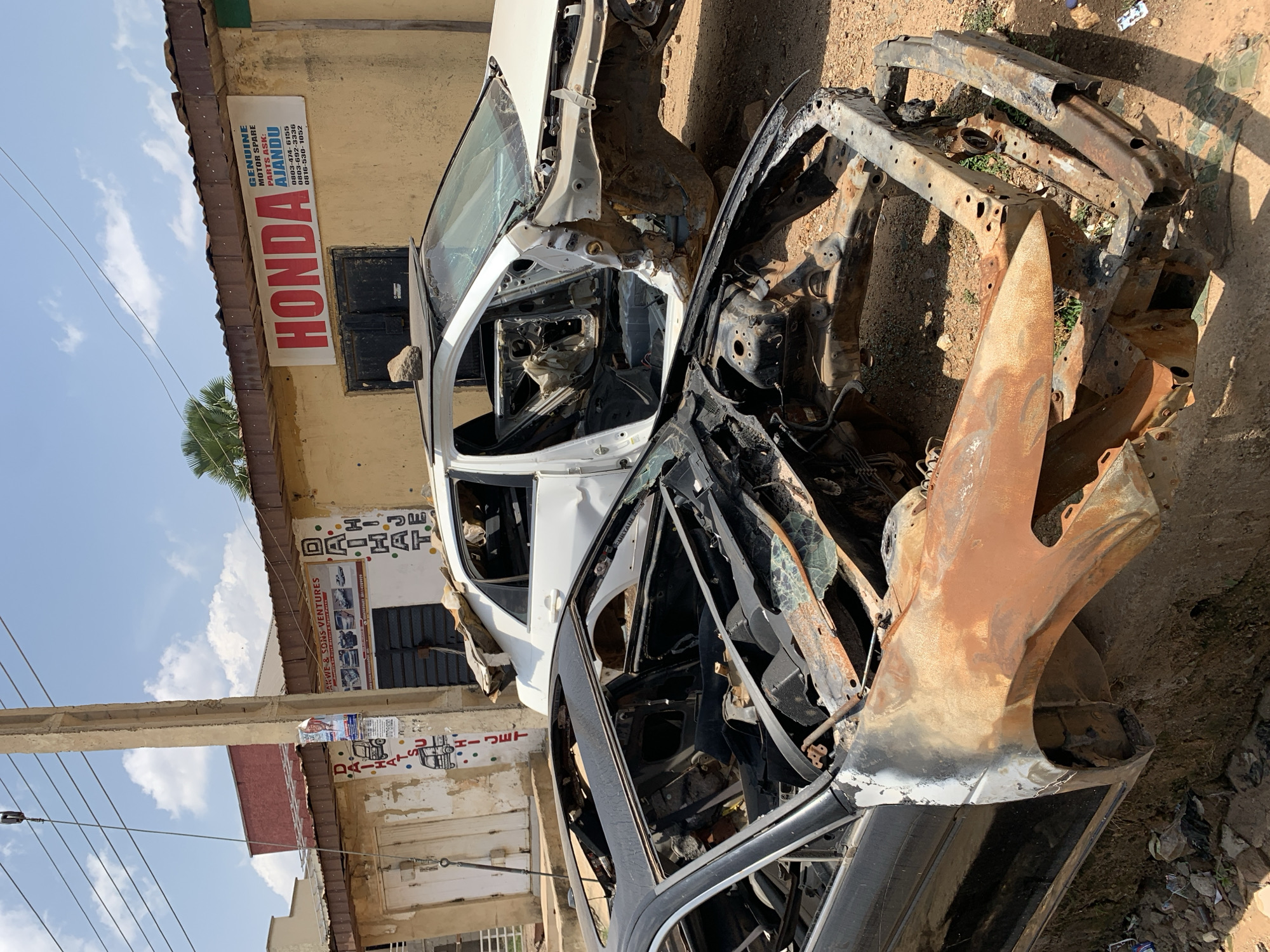 The image shows the aftermath of a vehicle fire in Rafin Par, Nigeria. The remains of at least two cars are visible; one is mostly white and relatively intact, while the other is heavily burned and rusted, with only the skeletal frame remaining. Both are situated on a dirt surface next to a building that is marked with various signage: a "HONDA" parts advertisement, as well as "DAIHATSU" advertisement. The sky is visible in the background. There are no people visible.