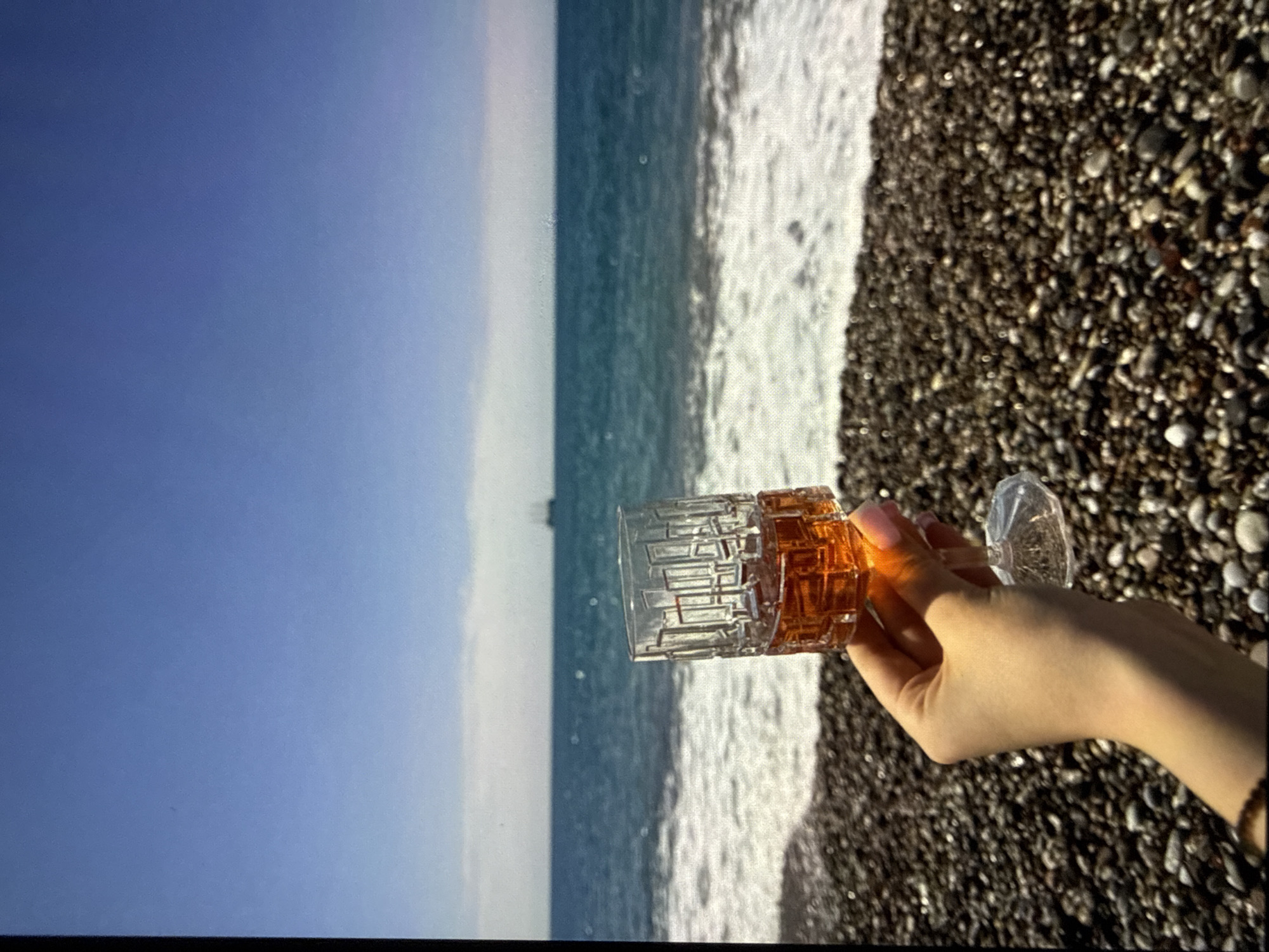 A hand holds a small glass of amber liquid on a pebble beach. The ocean and a clear blue sky are in the background, with a distant ship visible on the horizon. The scene is set on a beach in the city of Mykolaiv, Ukraine.