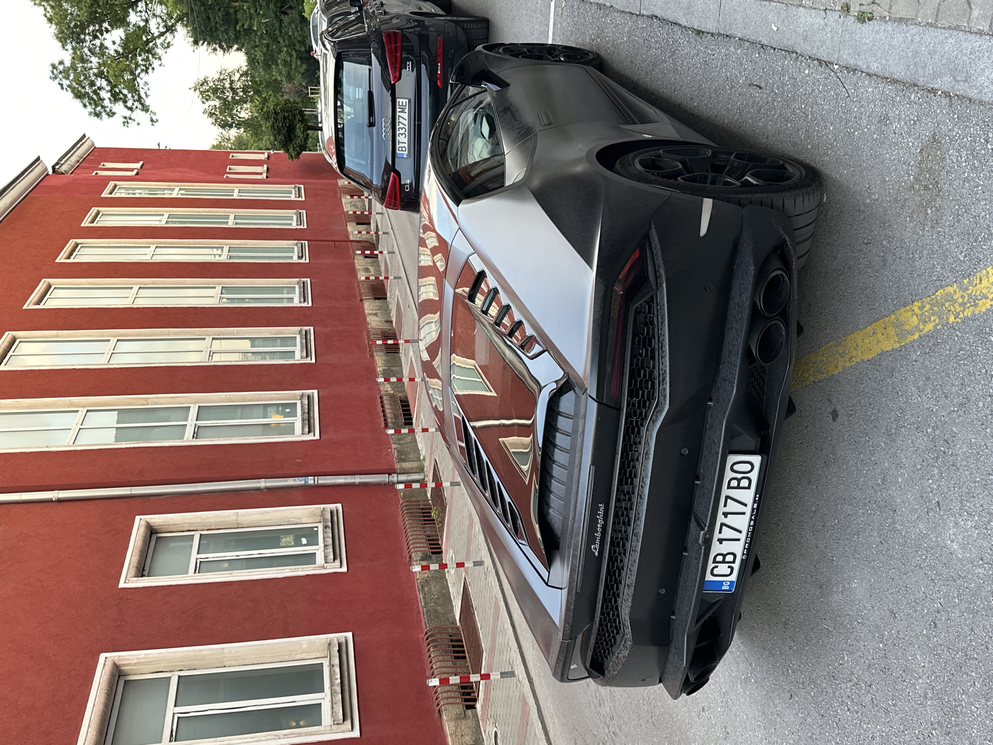 This media file features a striking, dark-colored Lamborghini sports car parked on a street next to a red building. The car's rear is prominently displayed, showcasing its aggressive design, large exhaust pipes, and a license plate that reads "CB 1717 BO BG". The building to the left has a repeating pattern of windows, some with a reflective quality that mirrors the surrounding environment. The street itself is paved with asphalt and marked with a yellow line, indicating a parking area.
A black Audi SUV is parked in front of the Lamborghini, and its license plate is partially visible, showing "BT 3377 ME". The scene appears to be set during the daytime, with overcast skies suggested by the ambient lighting. The setting is urban, with trees visible in the background, hinting at a pleasant, possibly temperate climate. The overall impression is one of modern luxury vehicles parked in a somewhat ordinary urban environment. The image is oriented vertically, suggesting it was taken from a slightly elevated position or that the subjects were captured from an unusual angle. There is no visible activity or people in the frame. The building's architecture and the street's appearance suggest a location in Eastern Europe, consistent with the provided location context of Veliko Tarnovo, Bulgaria.