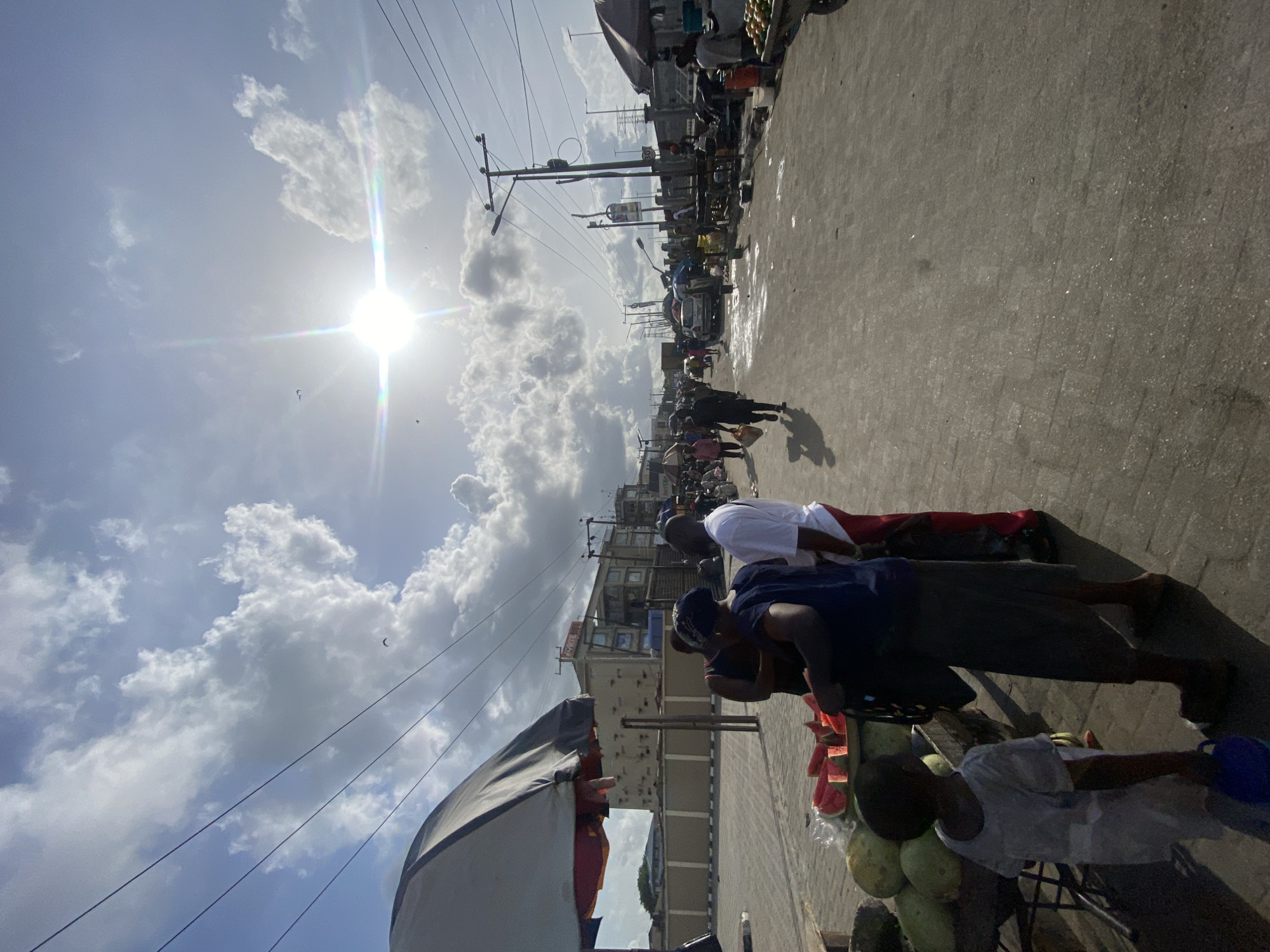 The image depicts a bustling street scene in Lagos, Nigeria. The sun is high in the sky, partially obscured by scattered clouds. Utility poles line the street, crisscrossed with wires. Along the right side of the image, a group of people are gathered around a vendor's cart, which is laden with watermelons and other produce. The vendor and customers are interacting around the cart. The road stretches out in perspective towards the horizon, lined with buildings, shops, and other vendors. The street is filled with people and vehicles.