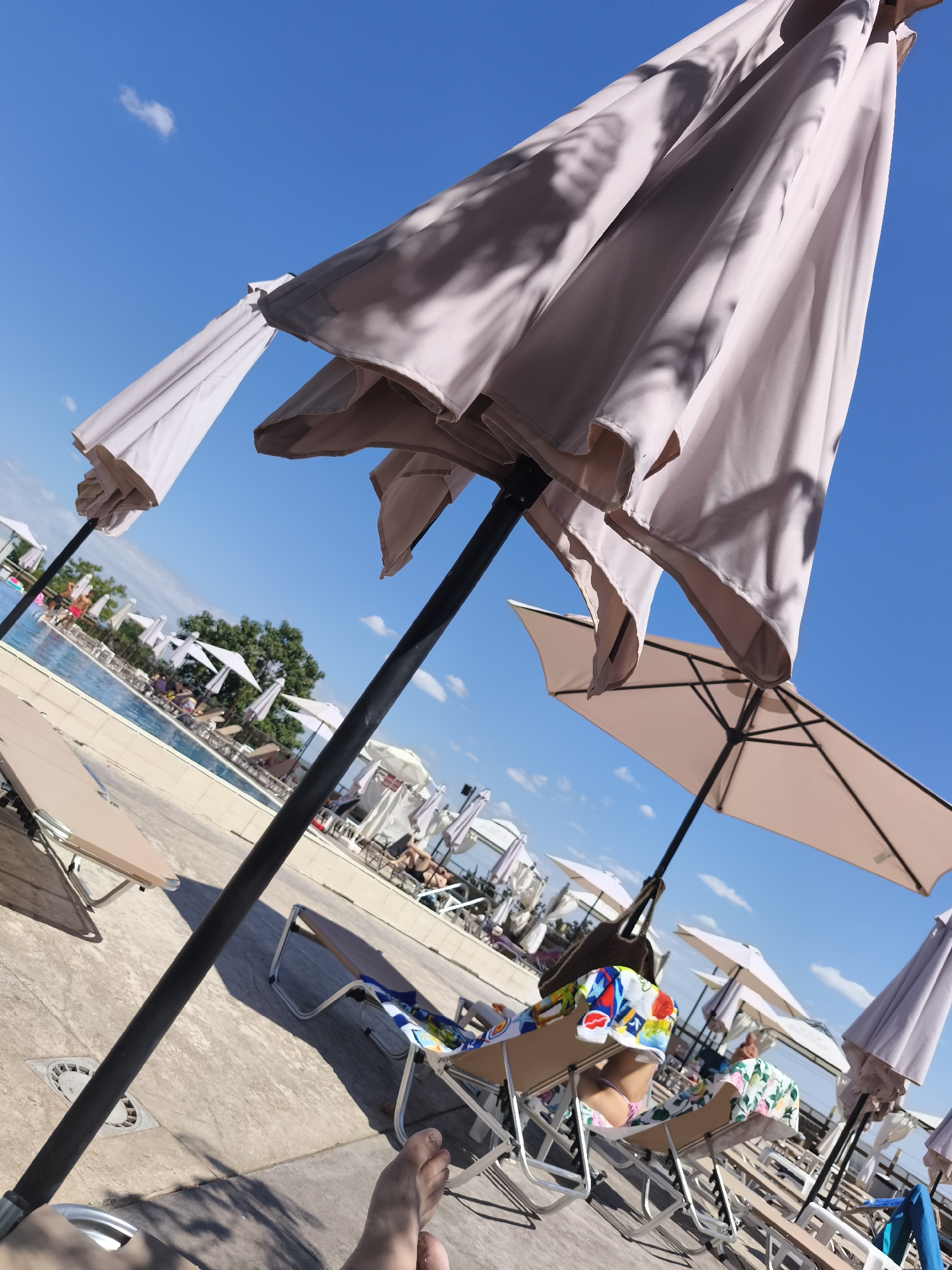 The image depicts a poolside scene on a clear, sunny day with a bright blue sky. In the foreground, a large, light-colored umbrella is tilted, casting shadows. Another open umbrella is visible to the right. Several empty lounge chairs are arranged on a paved area. In the middle ground, a swimming pool is visible, with a few people lounging on chairs and one person standing in the water. More lounge chairs and umbrellas are present around the pool area, with trees and foliage in the background. A person's feet are visible in the lower right corner of the image. The overall atmosphere suggests a relaxing resort or hotel environment.