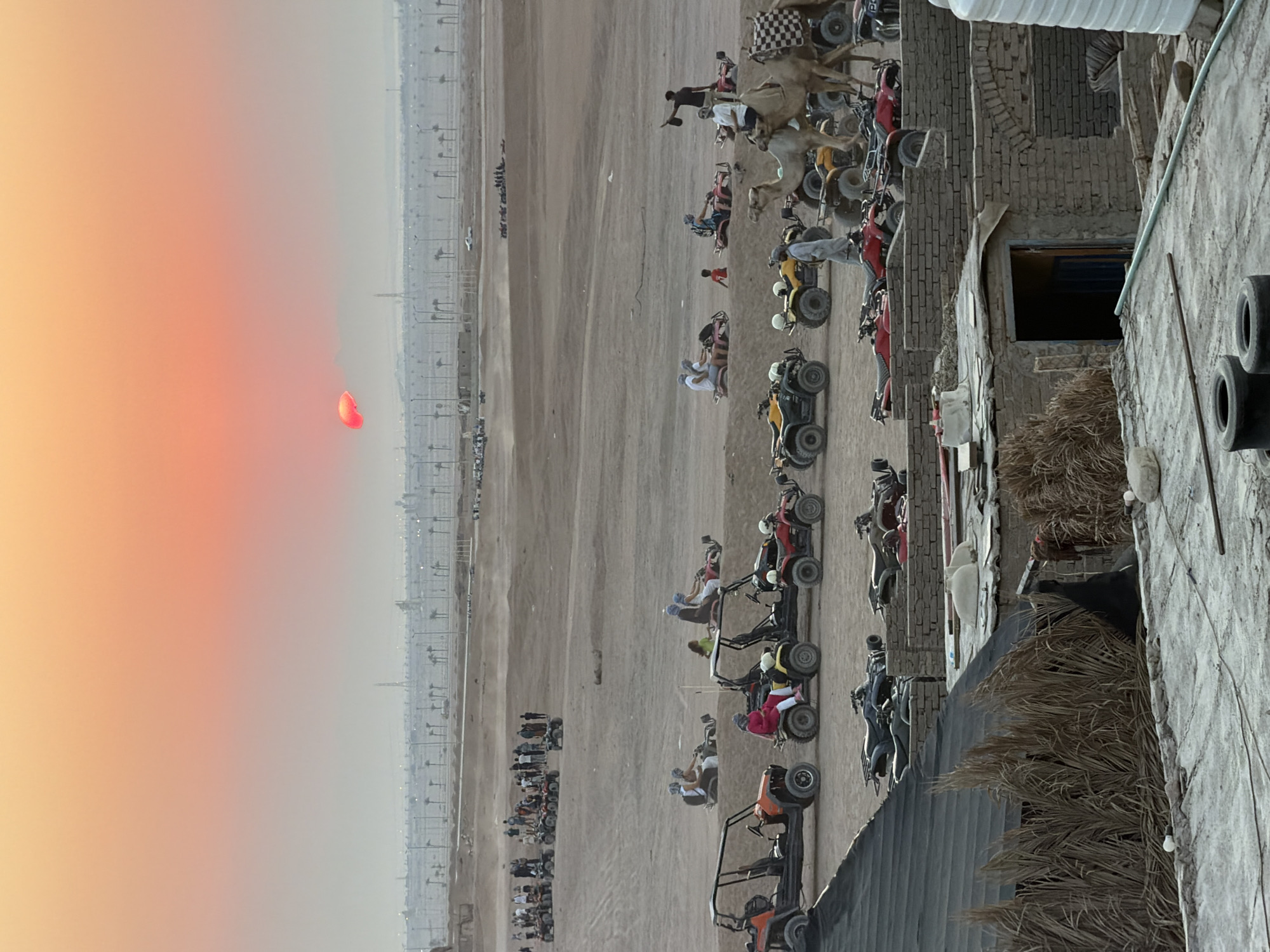 This is a full-frame, eye-level, medium shot photograph taken at dusk in Sharm El Sheikh, Egypt. The scene depicts a desert landscape dotted with various recreational vehicles and people. In the foreground, a straw-covered roof covers part of a simple, brick structure, with two tires visible on a concrete surface. Beyond this, in the mid-ground, several all-terrain vehicles (ATVs) and buggies are parked. Some are occupied by people, with individuals seated and ready for rides. Camel handlers stand near a group of camels, also prepared for tourist excursions. In the background, a line of palm trees and distant buildings suggests a developed area beyond the desert. The sky above is a gradient of orange and pink, indicating sunset, with the sun itself appearing as a small, reddish-orange orb partially obscured by atmospheric haze. The overall impression is of an organized desert adventure activity set against a picturesque twilight sky.
