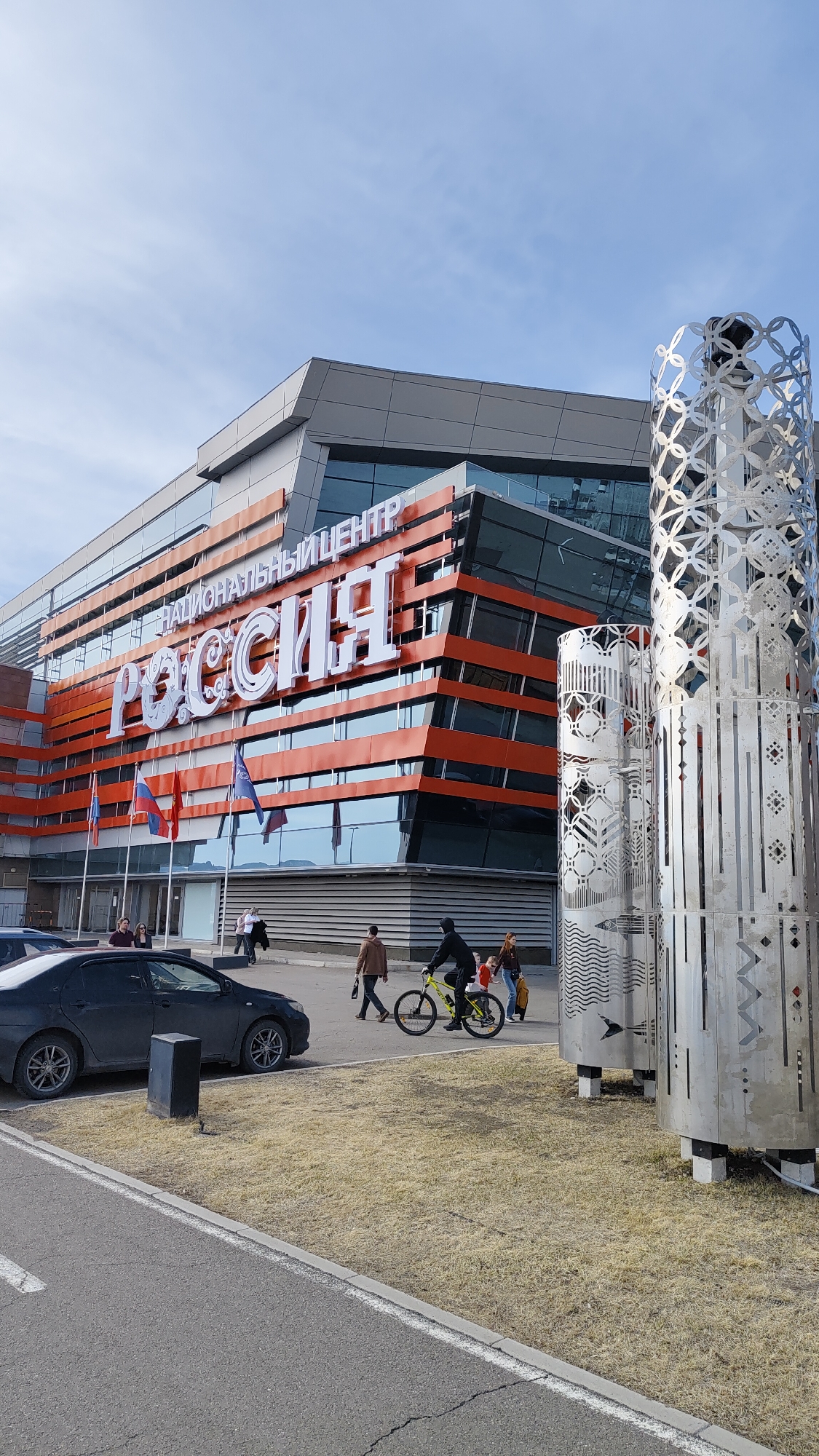 The image shows a building with "РОССИЯ" and "НАЦИОНАЛЬНЫЙ ЦЕНТР" written on its facade. Flags are displayed near the building's entrance. To the right of the building, there are metal structures resembling cylindrical towers with decorative patterns. A dark gray car is parked in the foreground on the left, near what appears to be a utility box. Several individuals are present in the scene: a man in a brown jacket is walking, and another person, dressed in black and riding a bright yellow bicycle, is accompanied by two other figures. The surrounding area includes a stretch of dry grass, a paved road, and a blue sky with some clouds visible in the background. The location is identified as Unknown, Unknown.