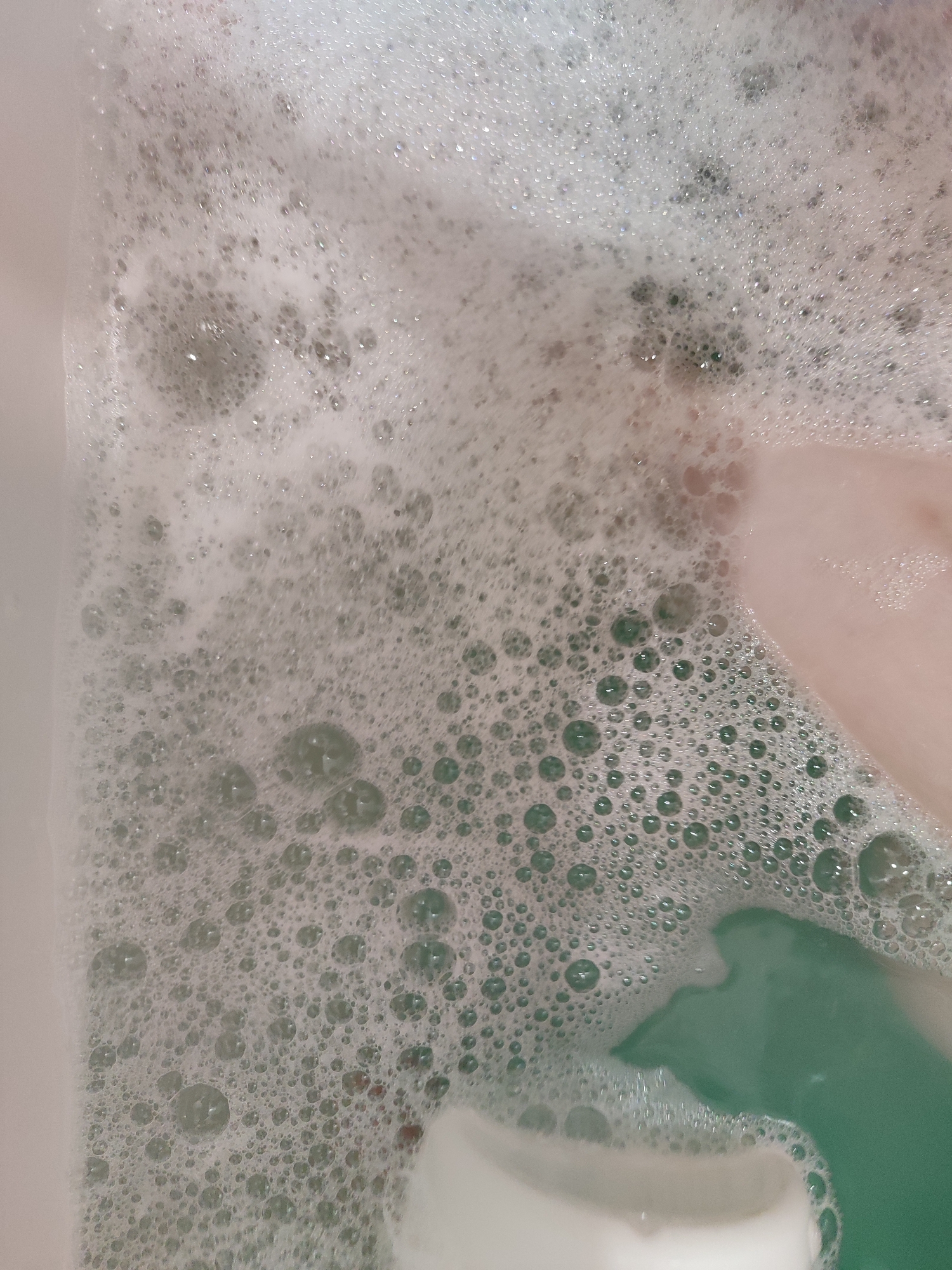The image presents a close-up, overhead view of a bathtub filled with water and a significant amount of white foam and bubbles. The scene depicts an activity of bathing.
The dominant feature is the dense, frothy layer of white bubbles that covers most of the water's surface. These bubbles vary in size, from tiny specks to larger, more defined spheres, particularly visible where the foam is thinner or closer to the edges. Beneath the foam, the water is a distinct light green or turquoise color, suggesting the use of a bath bomb or bath product.
On the right side of the image, parts of a human limb, likely a leg or foot, are visible, submerged under the foam and water. The skin tone appears light. In the lower right corner, a clearer patch of the green water is visible, alongside what appears to be a submerged green object or part of a bathing accessory, adding a deeper shade of green to the composition. The edge of a white bathtub, possibly made of porcelain, can be seen along the left side and bottom of the frame.
The setting is an indoor bathroom in Izhevsk, Russia. There are no specific cues within the image to indicate the time of day, weather, or emotions. No text is visible in the media file.
