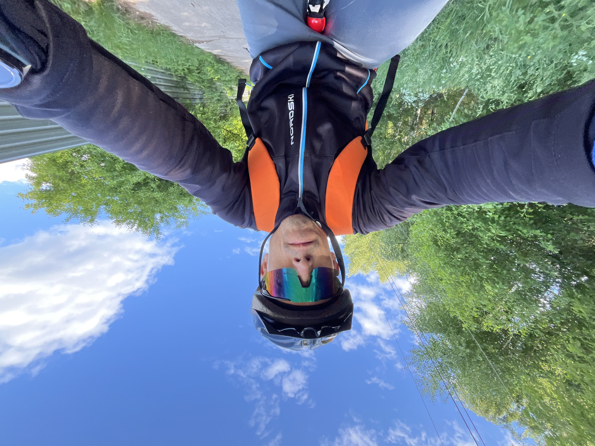 An outdoor selfie captures a person from a low-angle, looking up towards the sky, though the image is oriented vertically with the sky at the bottom. The central subject is an adult male, seen from the chest up, wearing a black cycling helmet and large, multi-colored reflective sunglasses. He is dressed in athletic wear, including a black long-sleeved shirt, and a black vest with distinct orange shoulder panels and light blue and white stripes down the front. The brand "NORDSKI" is visibly printed on the chest of the vest.
The man is taking the photograph, likely a selfie, possibly while cycling or during a break from the activity, given his attire. Above him (at the top of the image in its current orientation) are grey shorts or pants.
The background primarily features a bright blue sky with scattered white, fluffy clouds. Lush green trees and foliage are visible along the top edges of the frame, suggesting a natural or semi-urban outdoor environment. A narrow paved path or sidewalk runs along the top, next to grassy areas. Utility lines are also faintly visible against the sky and trees. A small puddle is present on the grass in one of the cropped views, indicating damp ground. The scene depicts a clear, bright daytime, suggesting good weather for outdoor activities in Chaikovsky, Russia.