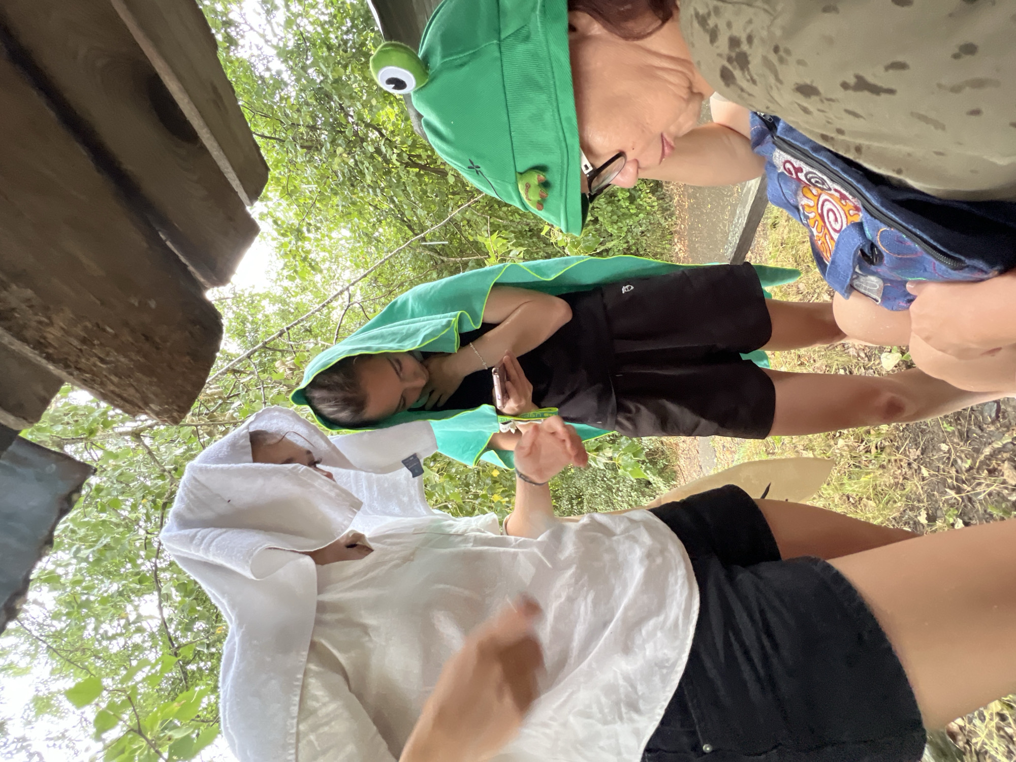 Three individuals are gathered under a wooden structure, possibly a lean-to or makeshift shelter, amidst lush green foliage. It appears to be raining, as evidenced by the water droplets visible on the ground and the towels draped over the heads of two people. The person on the left is wearing a white short-sleeved shirt and black shorts, with a white towel covering her head. Her left arm is extended, palm facing upwards, in a gesture that could be checking the rain or interacting with someone. The person in the middle is wearing a black t-shirt and black shorts, with a bright green towel draped over her head. She is looking down at a smartphone in her hand. The person on the right is wearing glasses and a green hat designed to look like a frog. She is looking downwards, with a blue bag with a sun motif resting on her lap. The overall environment suggests an outdoor setting, possibly in a park or rural area, and the presence of the wooden shelter indicates an attempt to seek protection from the weather. The background consists of trees and vegetation. The scene does not contain any identifiable landmarks or specific features that would allow for pinpointing a location within the city of Sallanches, France, or anywhere else.