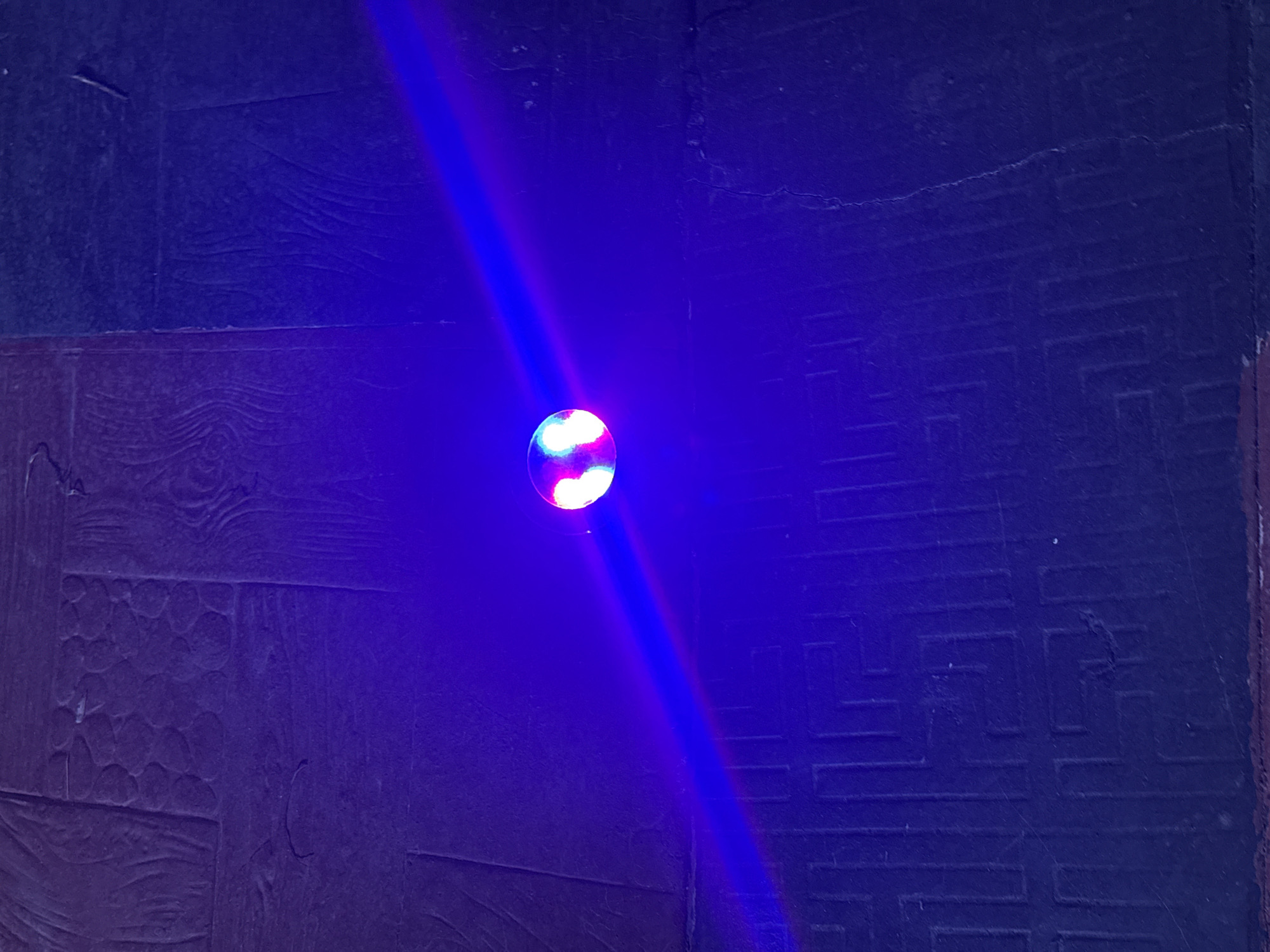 In the city of Abuja, Nigeria, a low-angle shot captures a close-up view of a circular object, possibly a light fixture or a toy, emitting a vibrant blue laser beam. The object, centrally located and slightly to the left, displays a split of red and green light. The laser beam extends diagonally across the frame, illuminating a textured floor that appears to be made of dark stone or concrete tiles. The floor exhibits a patterned relief, with rectangular sections containing embedded circular shapes and linear grooves. The overall ambiance is dark, with the intense blue light of the laser dominating the scene, creating a dramatic and slightly abstract effect.