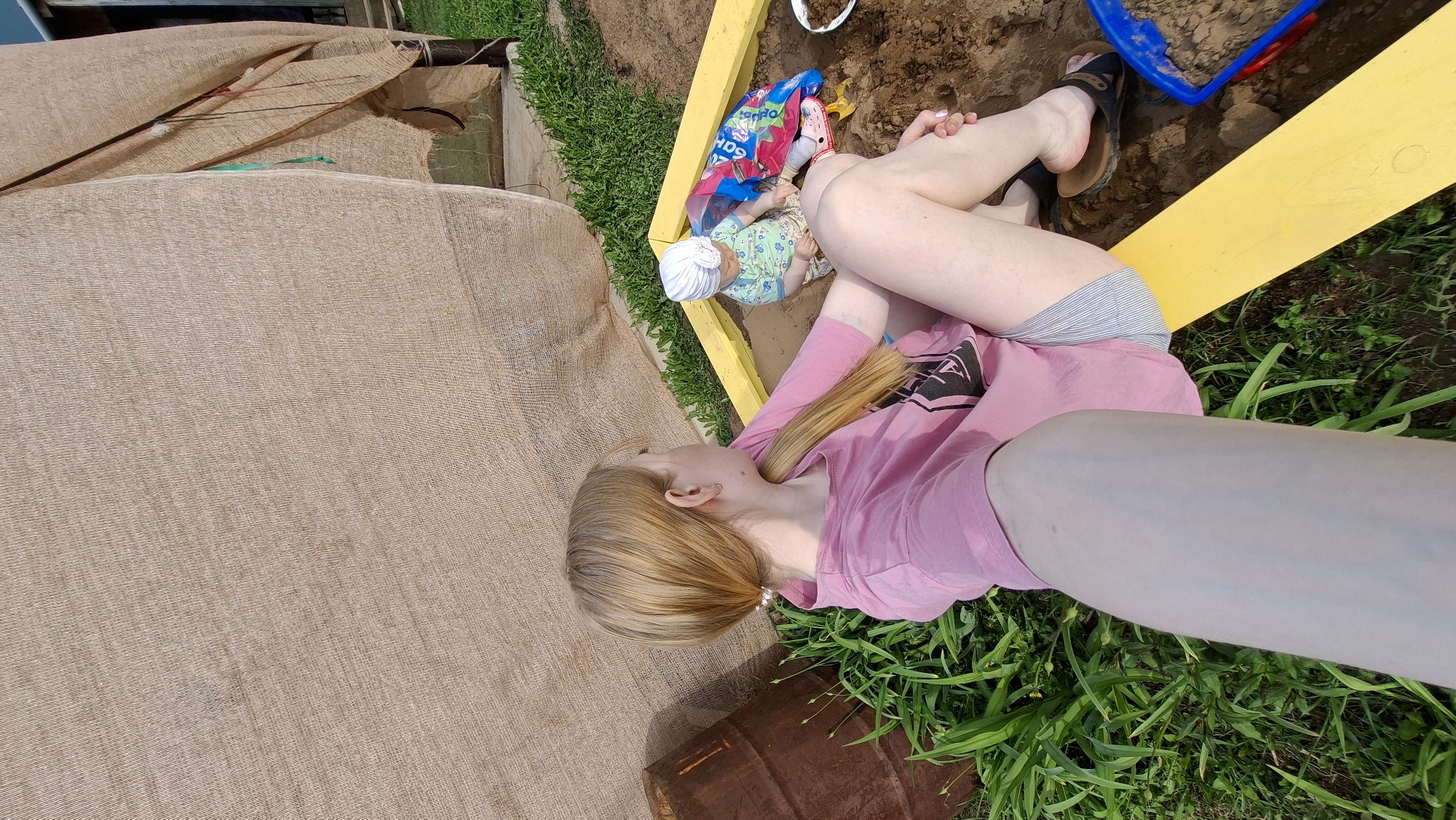 The image presents an outdoor scene in what appears to be a yard. In the upper-right quadrant, there is a child's sandbox with sand, a blue toy, and a yellow border constructed from wooden planks. Adjacent to the sandbox, a woman is seen lying on the grass, wearing a pink top and grey shorts, with her legs positioned partially within the sandbox area. Next to her inside the sandbox lies what appears to be a doll.
To the left of the woman, there is a large, light-brown woven material covering an area, possibly a garden bed. In the bottom center, there is a rusty barrel partially obscured by grass. The environment includes green grass covering the ground surrounding the sandbox and barrel.
The location is specified as Большой Букор, Russia.