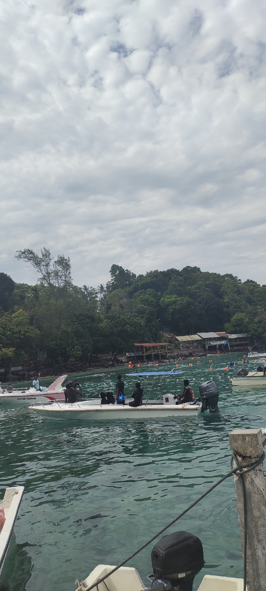 This is a vertical, medium-distance, eye-level shot taken from a boat in the waters of Sabang, Indonesia. The foreground features the rear of a white boat with a black outboard motor. The water is calm with visible ripples, and it's a translucent green color, revealing the sandy bottom in places. In the mid-ground, several other boats are present, some with people on board. One prominent white boat has at least four individuals, some seated and some standing. Another white and pink boat is to the left, also with occupants.
Beyond the boats, the coastline is lined with lush, green, tropical vegetation, indicating a jungle or forest. Several buildings, possibly small shops or huts, are visible nestled amongst the trees. The water is populated with many people wearing orange life vests, some swimming and others in smaller floating devices. The sky above is overcast with large, fluffy, white clouds. The overall scene depicts a popular tourist area, likely a beach or snorkeling spot, within Sabang.
