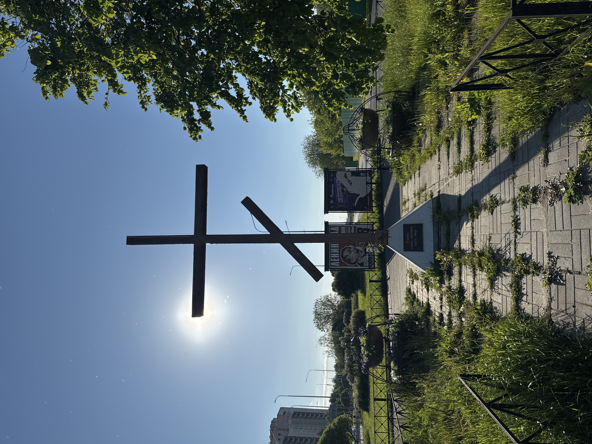 The image depicts a sunny outdoor scene in Saint Petersburg, Russia, featuring a large wooden Orthodox cross in the foreground. The sun is positioned directly behind the cross, creating a silhouette effect. Behind the cross, there is a memorial with posters and flowers, surrounded by a metal fence and overgrown grass. A paved walkway leads toward the memorial. In the background, there are buildings, trees, and streetlights. The sky is blue with scattered leaves visible at the top of the frame.