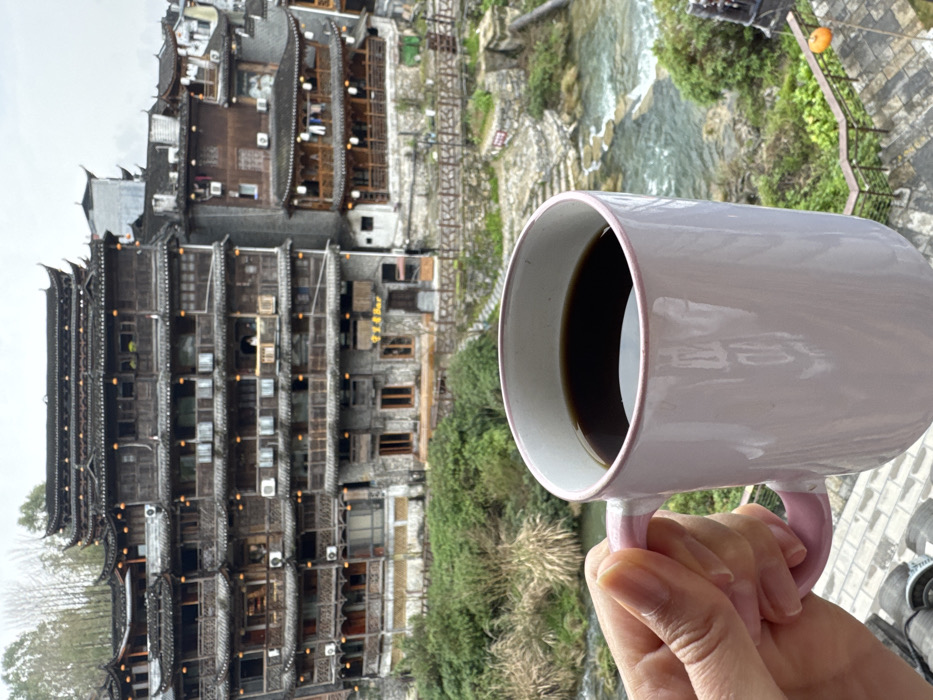 This image presents a detailed view of the traditional riverside architecture in the city of Furong, China, under an overcast sky. In the immediate foreground, a person's hand, exhibiting a medium-dark skin tone, firmly grips the light pink handle of a light pink ceramic mug. The mug contains a dark liquid, consistent with coffee, and features a subtle darker pink rim.

The perspective is from an elevated vantage point, looking down into a valley. Below the mug, a lush, steep hillside covered in various green and light brown vegetation slopes towards a clear river with visible cascades and flowing water. A dark wooden railing runs along the riverbank.

The midground and background are dominated by a complex of multi-story traditional Chinese wooden buildings, characteristic of the region, built directly into the steep terrain. The most prominent structure, located left of center, is a large, dark wooden building appearing to be at least five to six stories tall. It features numerous balconies with intricate wooden railings, multiple windows, and visible outdoor air conditioning units on several levels. Its roof structure consists of multiple tiers with traditional upturned eaves. At its base, a sign with Chinese characters and the English word "Bar" is affixed. To the right, additional traditional buildings with similar architectural styles extend along the river, one of which shows clothes hanging to dry on an open balcony. The overall scene depicts a blend of historical architecture and natural landscape.