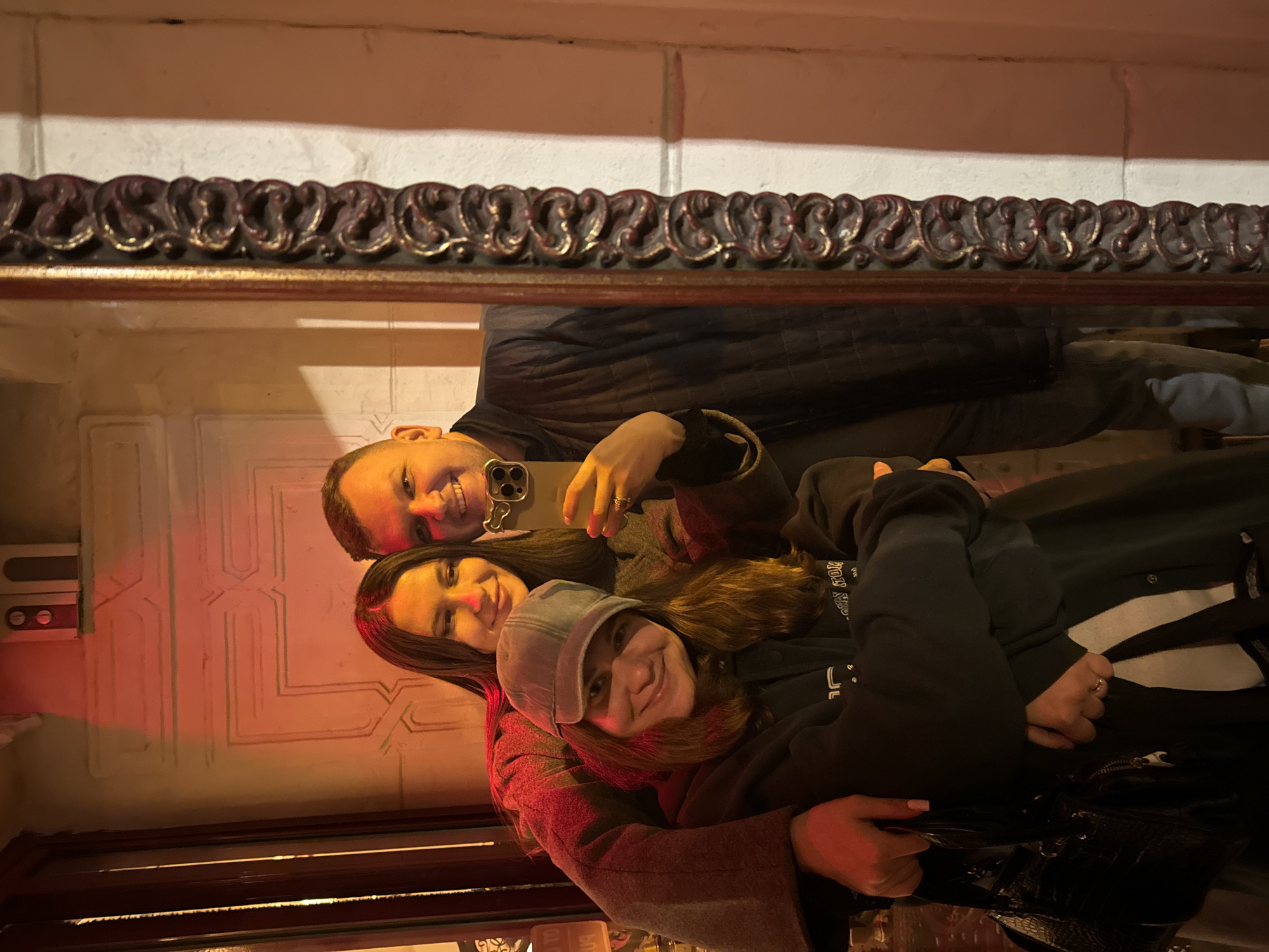 Three individuals are reflected in a mirror with an ornate, dark-colored frame. The reflection shows the individuals taking a selfie. The person on the left has short hair and is smiling, holding a phone to capture the image. A woman is positioned in the middle, also smiling. The person on the right wears a gray cap and a dark hoodie. The reflection also shows a patterned wall in the background, lit with a red glow. A small, rectangular box is attached to the wall. The scene seems to be indoors. It is impossible to verify the city and country from the given visual data.