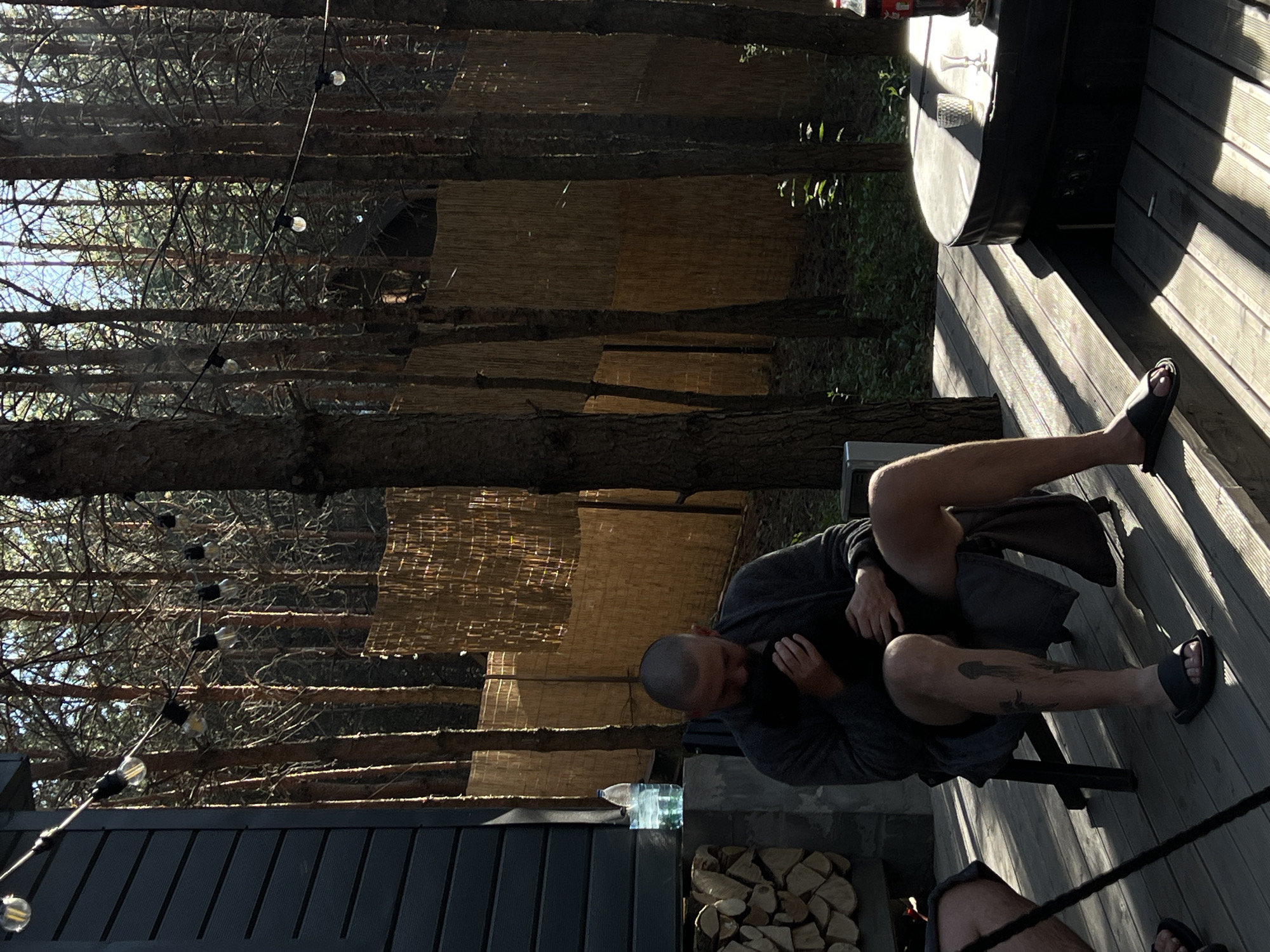 In the village of Ivachiv Horishnii, Ukraine, a man with a shaved head sits on a chair on a wooden deck, leaning forward and holding something to his chest. He is wearing a gray robe and shorts. His left leg is visible, showing a tattoo on his calf. To his left, a stack of chopped firewood is visible next to a dark-sided building. Behind the man, a bamboo fence stretches across the scene, with tall trees behind it. String lights with exposed bulbs are visible in the upper portion of the image, spanning across the trees. To the right of the man, a dark-colored circular object, possibly a hot tub, is present on the deck, with two glasses and a bottle of Coca-Cola on its surface. The lighting suggests it is daytime, with strong shadows cast by the sun.