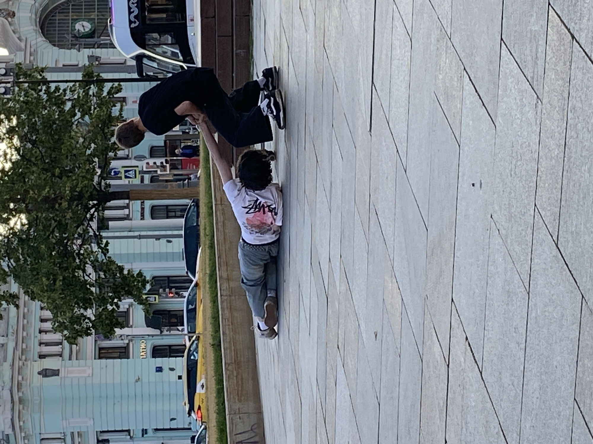 In a city square paved with large, gray stone tiles, a person lies prone on the ground, facing downwards, with their arms extended forward. A second person stands behind them, holding the prone person's left hand and wrist, and appears to be pulling them up. The prone person wears a white t-shirt with a graphic design and light-colored pants, and is barefoot. The standing person wears a black t-shirt, black pants, and black sneakers. In the background, a light blue building with arched windows and architectural details can be seen, along with a green tree and yellow taxis.