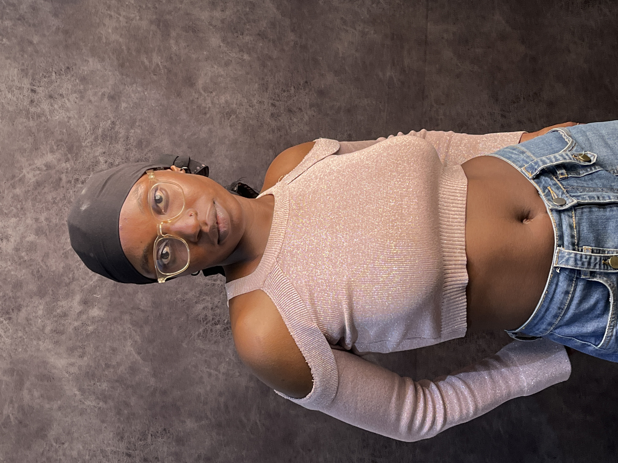 A person is lying on a mottled grey surface. They are wearing a black head covering, glasses with gold frames, a long-sleeved light pink cropped top with cutouts at the shoulders, and blue denim shorts with buckles. The shorts are worn low enough to reveal the person's abdomen. The location is not specified as Jos, Nigeria.