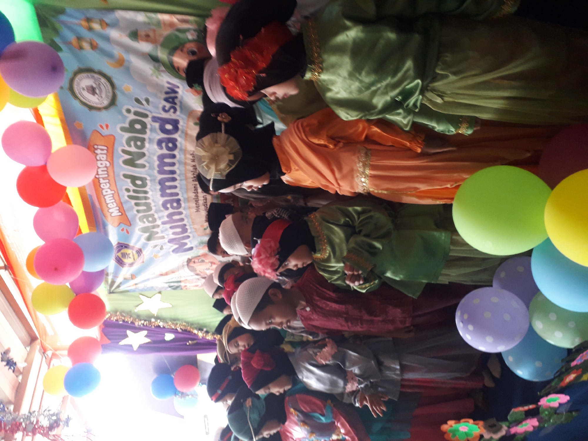 In Cankuang, Indonesia, a group of children are gathered for a Maulid Nabi Muhammad SAW celebration. The children, dressed in traditional attire, stand in rows, with many wearing hijabs and some adorned with decorative headpieces. The backdrop features a banner with Islamic calligraphy and imagery, including a mosque and crescent moon, suggesting a religious event. Colorful balloons are scattered throughout the scene, adding to the festive atmosphere. The event appears to be taking place indoors, possibly in a community hall or school, with decorations and lighting indicating a planned celebration.