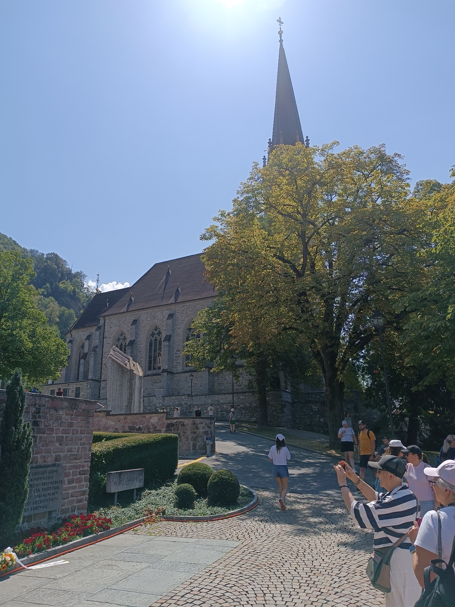 The image captures a sunny day scene outside a stone church in Vaduz, Liechtenstein. A prominent steeple with a cross on top rises above the church's roof, partially obscured by the lush green foliage of a large tree in the foreground. Several people are visible in the frame, some walking and others standing. One individual, wearing a striped shirt and cap, is actively taking a photograph with a phone. Another person in the foreground, wearing a white top and denim shorts, is walking away from the camera. The church itself is constructed of large stone blocks with arched windows. A paved pathway leads up to the church, bordered by a low stone wall and meticulously trimmed hedges. A stone memorial plaque is affixed to the wall, and a small cluster of red flowers is planted nearby. In the background, a tree-covered hillside slopes upwards, characteristic of the mountainous landscape surrounding Vaduz. The sky is clear and bright blue, with the sun prominently visible, creating lens flare in the upper right portion of the image.