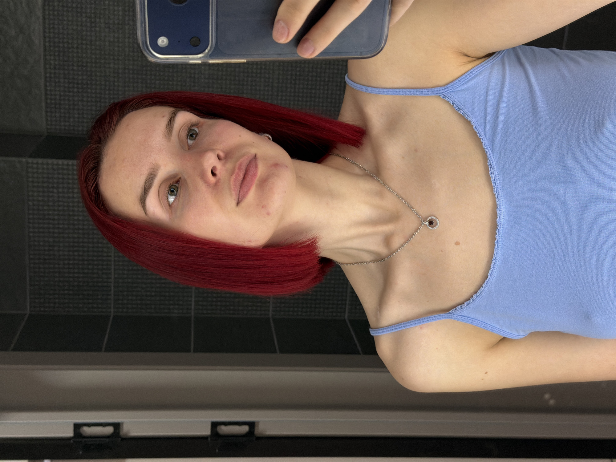 The image, rotated 90 degrees counter-clockwise, depicts a young woman taking a selfie in an indoor setting, likely in Chaikovsky, Russia. She has bright, short red hair styled in a bob cut and light-colored eyes that gaze directly at the camera. Her eyebrows are distinct, and her lips are slightly parted. Visible on her face are some blemishes, particularly on her chin and forehead.
She is wearing a light blue spaghetti strap camisole top and a silver chain necklace with a circular pendant that contains a dark red or brown stone. Her left arm, holding the smartphone, is visible at the top of the frame, showing the phone's dark blue case with a clear transparent cover. The background features dark gray or black tiled walls with a subtle grid pattern, and a horizontal surface, possibly a shelf or vanity, below her. Her expression is neutral and calm. No visible text is present in the image.