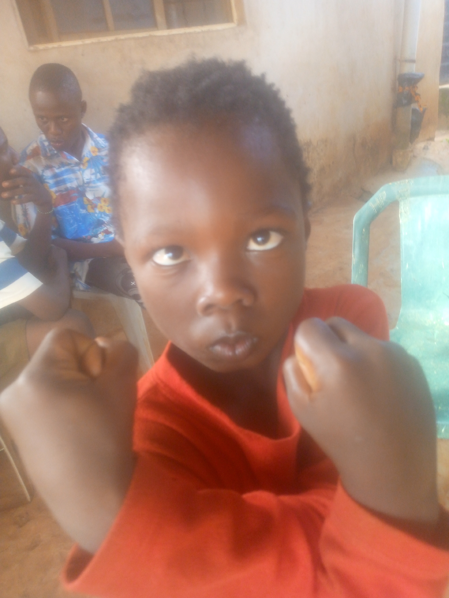 A young child with brown skin, wearing a red t-shirt, is the primary subject of the image. The child is holding both fists up near their shoulders. The background is a wall with a window and a person with a patterned shirt. Another person's arm and shoulder are visible. A light green plastic chair is visible to the right of the child. The location is Unknown, Unknown.