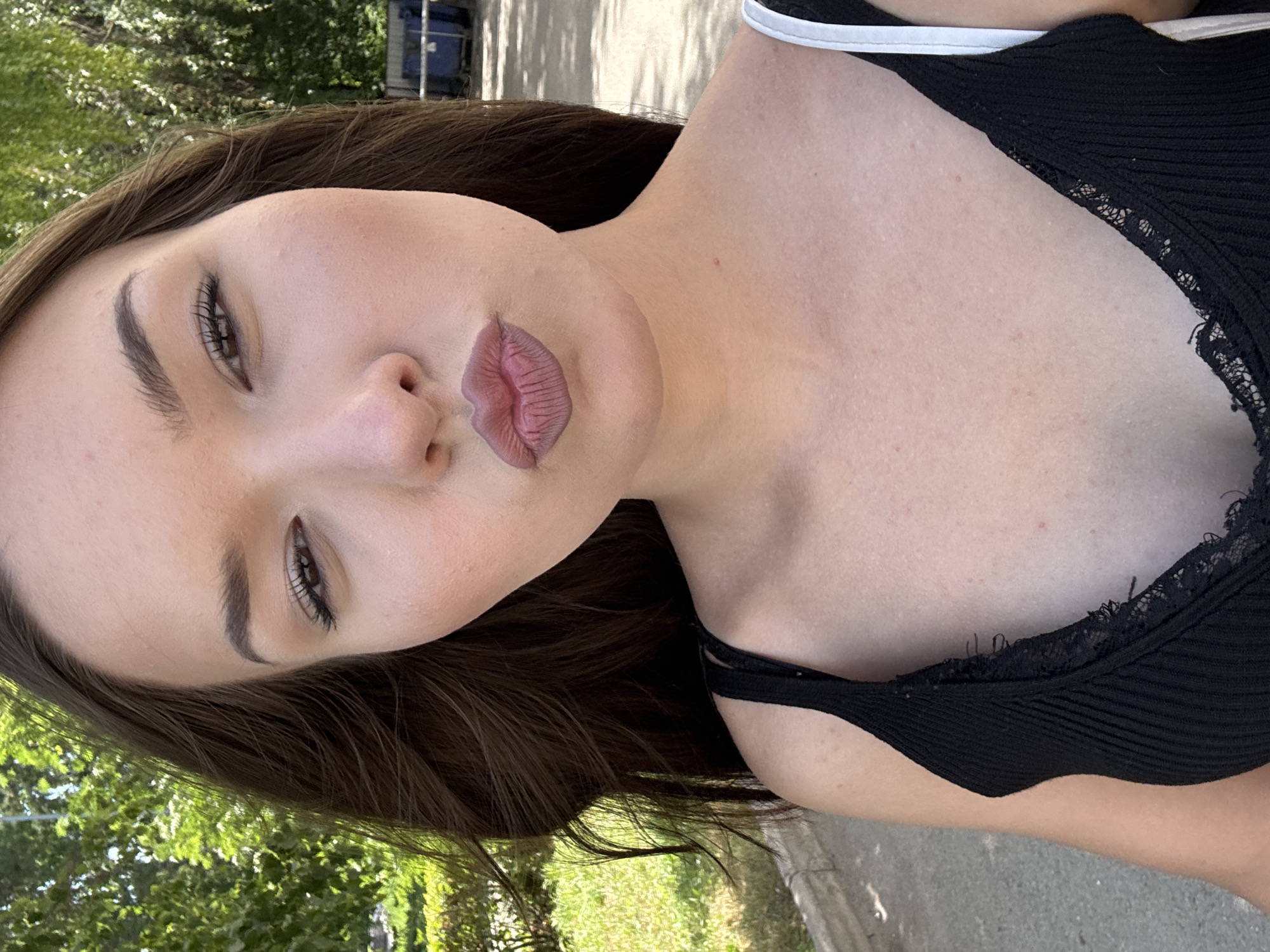 The image shows a close-up selfie of a young woman with dark brown hair. She is wearing a black tank top with lace trim. Her lips are pursed, and she is looking towards the camera with her right eye slightly closed. The background appears to be an outdoor setting, possibly a street or park, with green trees and foliage visible. There are also some blue bins in the background, suggesting an urban or suburban environment. There is no information within the image that specifies the location as Belovo, Russia.