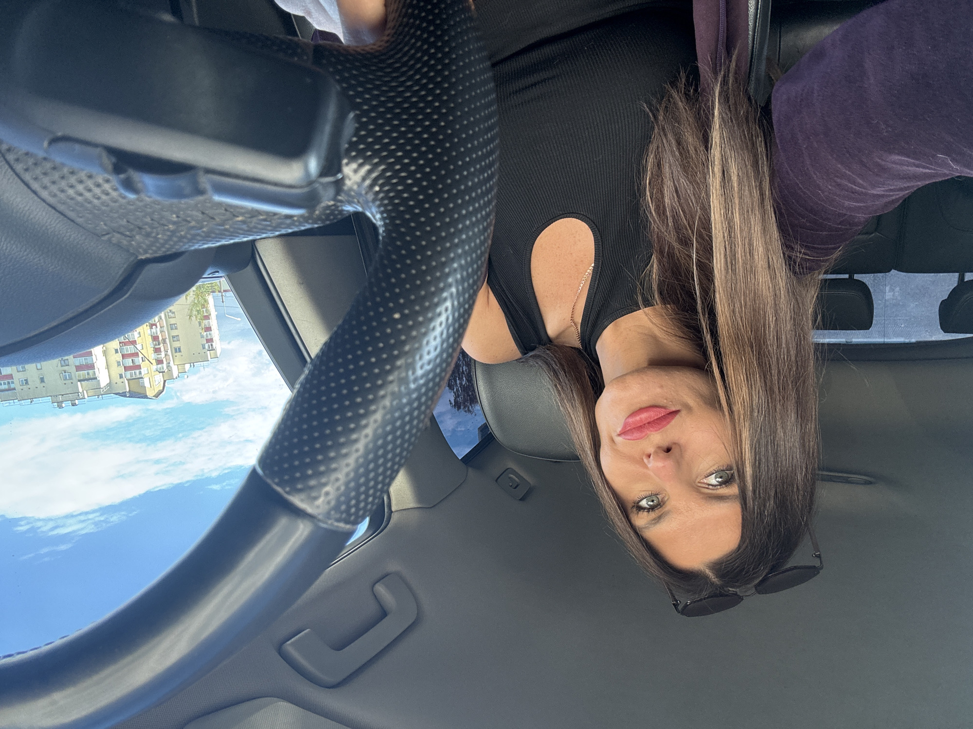 The image shows a woman sitting in the driver's seat of a car, taking a selfie. The photo is oriented sideways. She has long, brown hair, green eyes, and is wearing a black, ribbed dress with a cutout neckline, and a purple hooded sweatshirt. Sunglasses are resting on her head. The car's steering wheel is partially visible in the foreground. Outside the car, through the windshield, a bright blue sky with scattered clouds is visible, along with apartment buildings and trees. The sun appears to be shining, suggesting it is daytime. The setting appears to be an urban environment in Chaikovsky, Russia. The woman has a slight smile and looks directly at the camera.