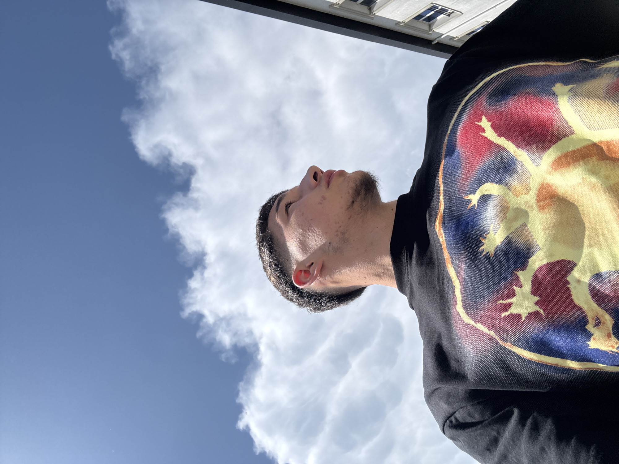 The image shows a low-angle view of a person looking up at a partly cloudy sky. The subject is a young man with short dark hair and stubble, wearing a black shirt with a large, colorful print. Above him are white clouds against a blue sky, and a building is partially visible at the top of the frame.
The specific location in Zlatograd, Bulgaria, cannot be determined based solely on this image. More information is needed to pinpoint the exact spot.
