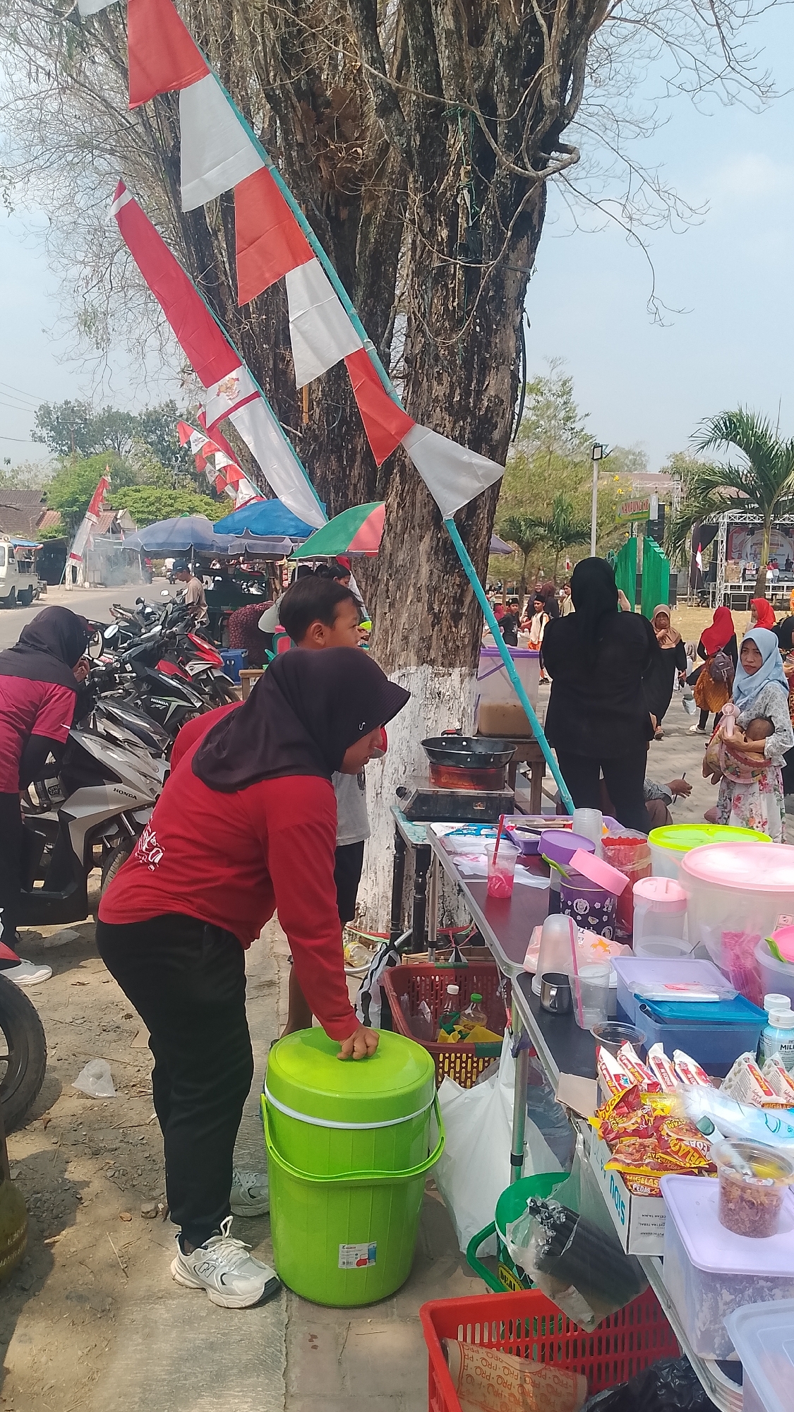 This is an outdoor, eye-level, medium shot of a market scene, likely in Karanglangu, Indonesia, judging by the flags which appear to be Indonesian flags, red and white stripes on poles. The foreground features a variety of food stalls and a crowd of people.
In the immediate foreground, to the left, is a large, bright green plastic cooler. A person wearing a red long-sleeved shirt and black pants is leaning over it, with their right hand on the lid. Their face is not visible. To the left of the cooler, a portion of a motorcycle's rear wheel and tire is visible.
A table with various items is set up in the middle of the frame. On the table, there are several containers of food, plastic cups, and packets of snacks. A black frying pan is visible on a portable stove behind the table. In the background, more market stalls are visible, some with umbrellas providing shade. There are several motorcycles parked behind some stalls.
People are milling about the market. To the left, two women wearing hijabs are interacting near the parked motorcycles. One is wearing a pink shirt and the other a dark colored top. In the center background, several people are walking around. To the right, a woman wearing a light blue hijab is holding a baby. Further back, a stage or performance area is visible with palm trees and some structures. The sky is bright and clear, suggesting a sunny day. The ground is paved with what appears to be asphalt or concrete.